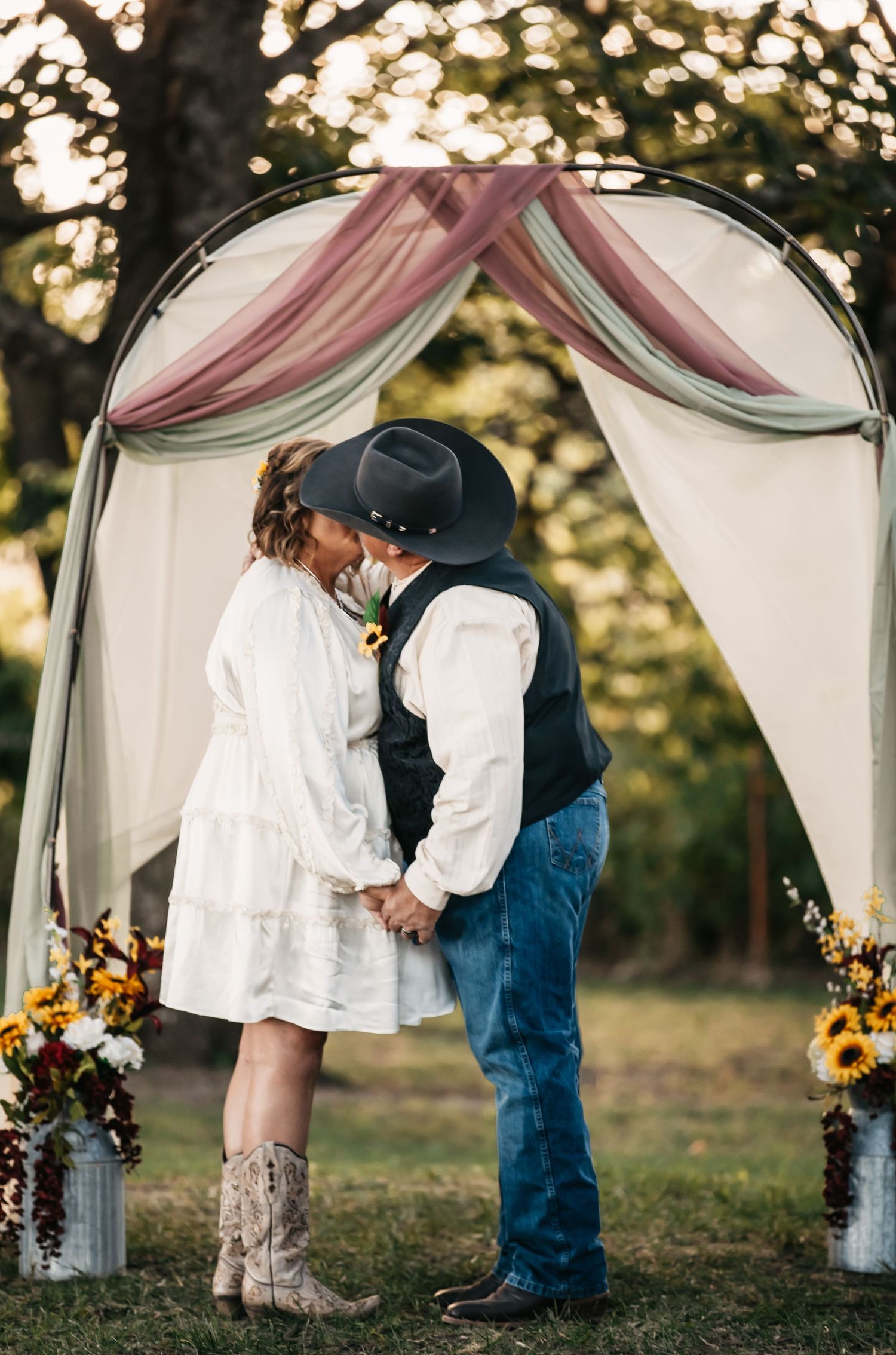 Romantic moment under white draped wedding arch decorated with sunflowers at sunset.