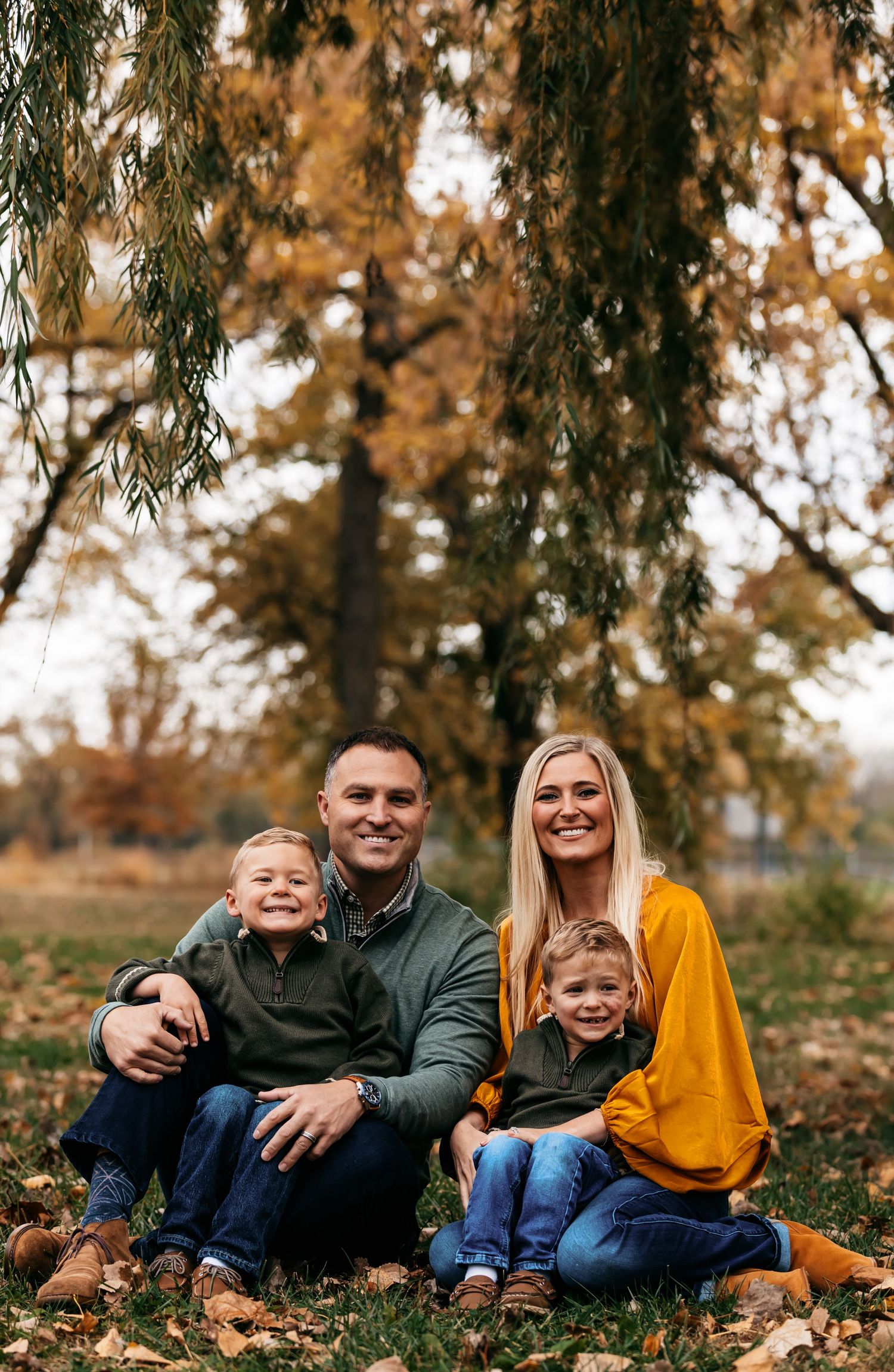 Family relaxing together on fallen autumn leaves under willow trees.