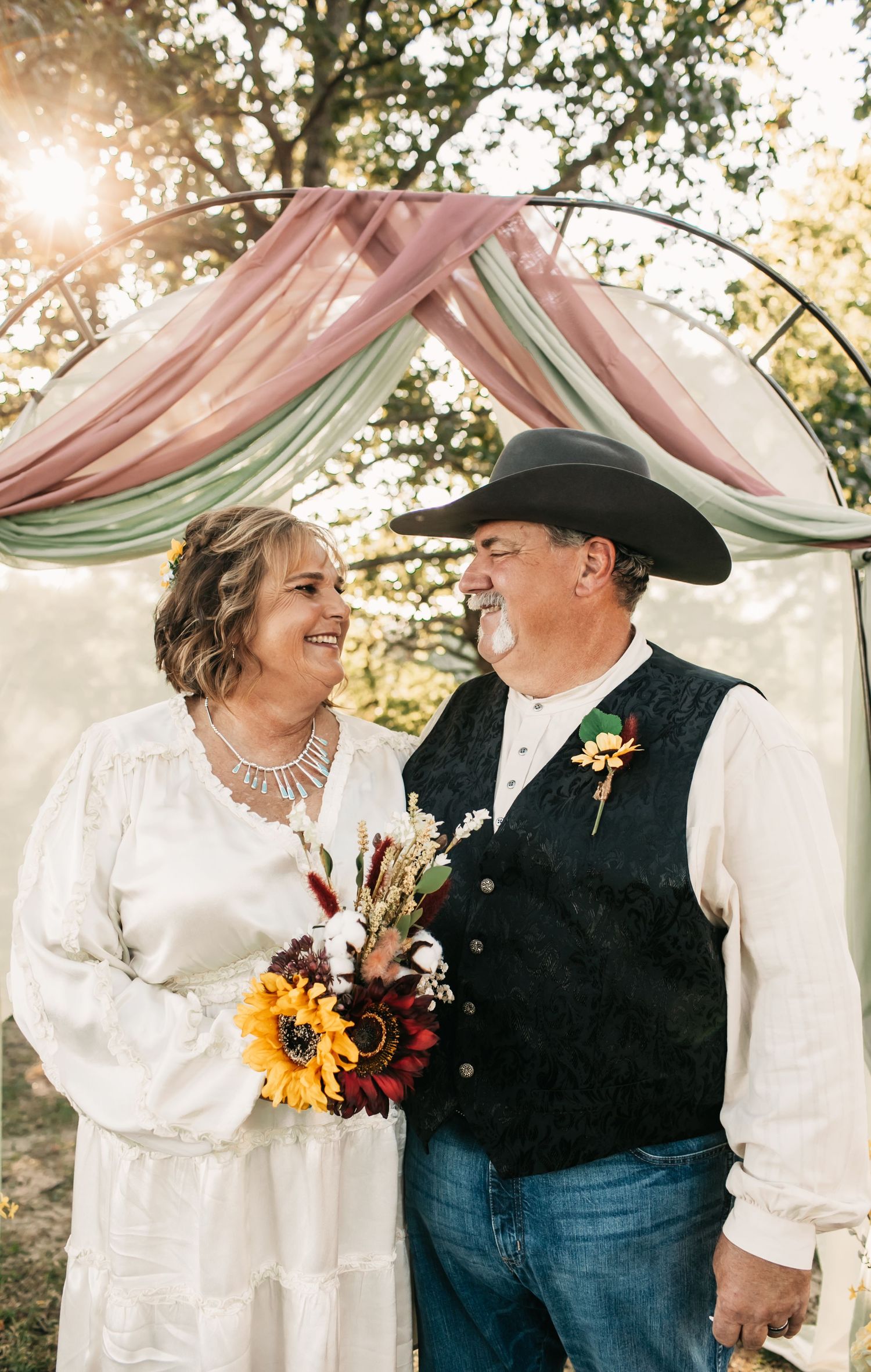 Couple sharing intimate moment under wedding arch with sunflowers and western attire.