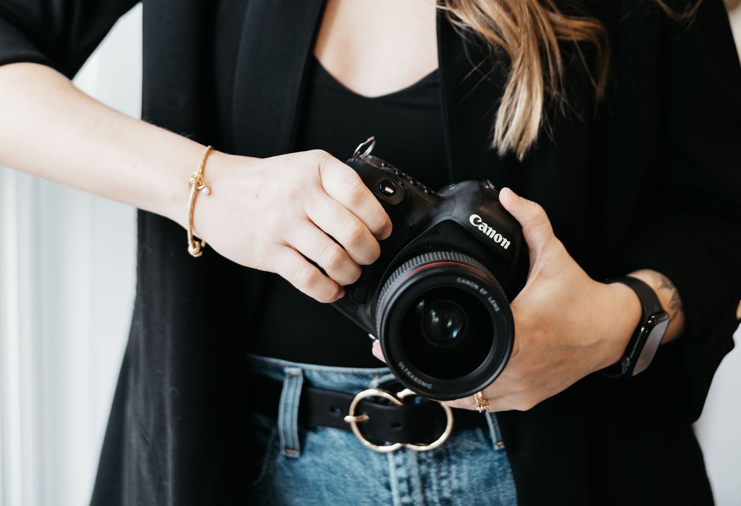 Photographer holding a Canon DSLR camera in a black outfit against a light background.