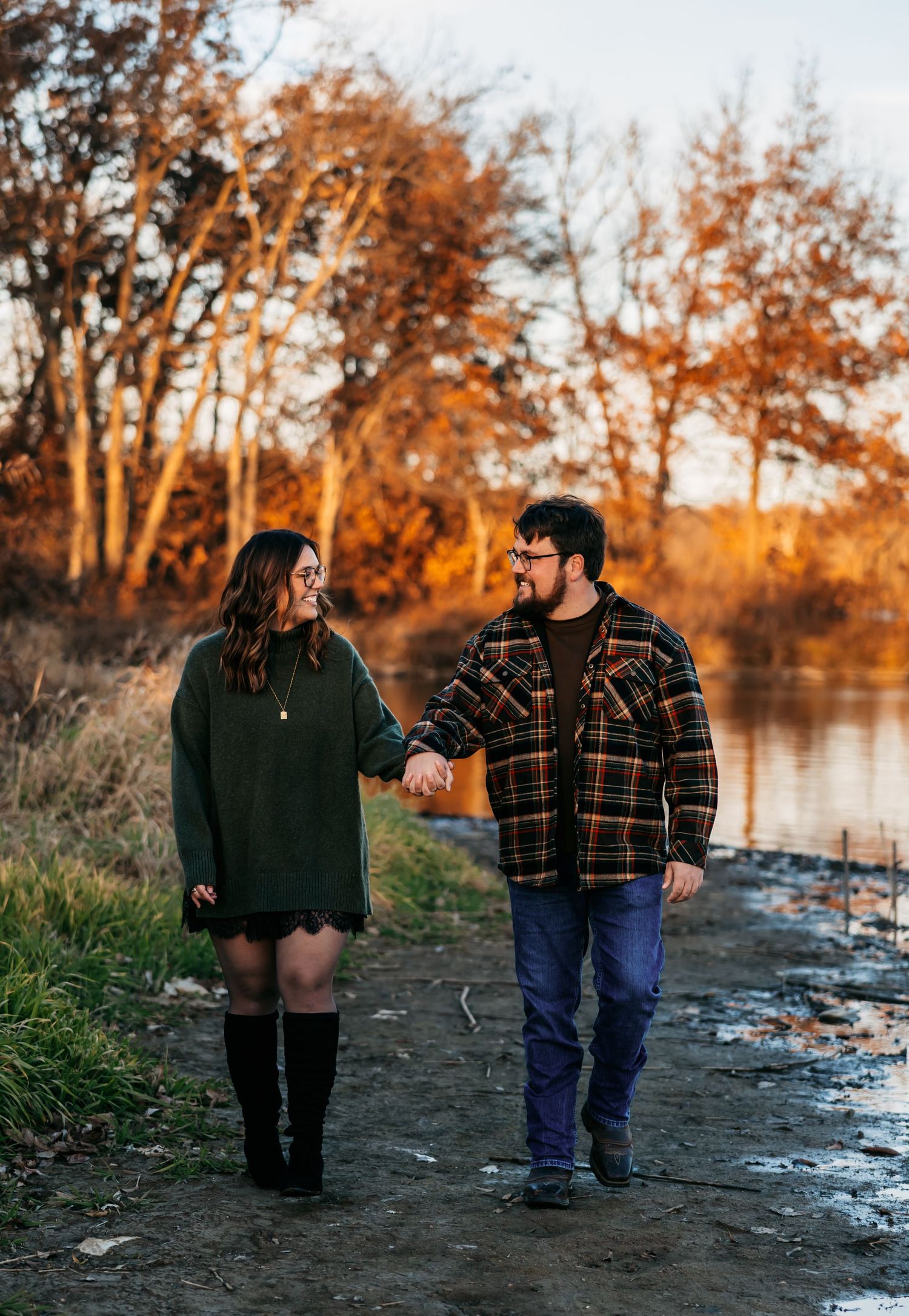 A couple walks together along a riverbank during golden hour wearing casual fall attire.