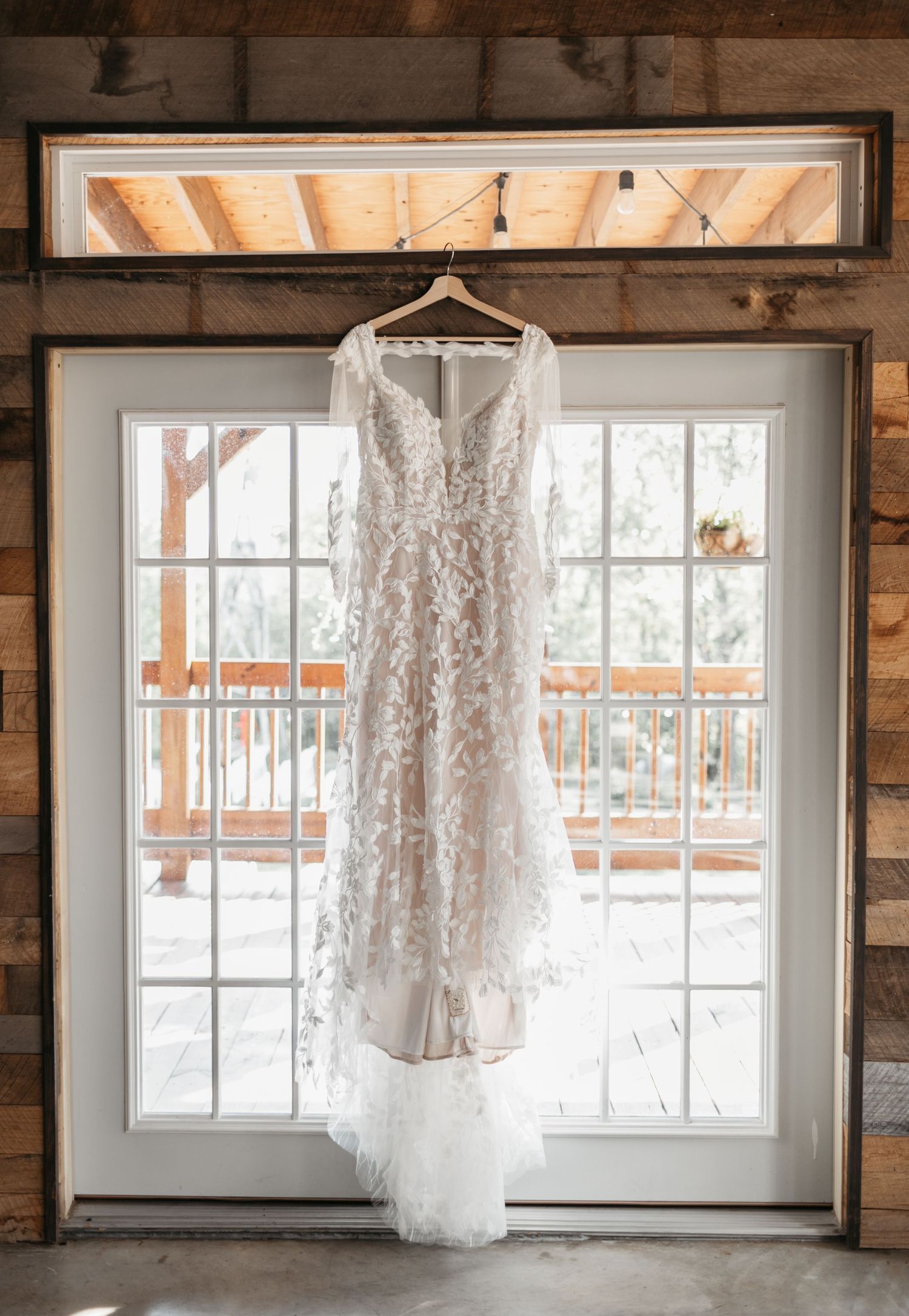 White lace wedding dress hanging in front of bright windows in a rustic wooden room.