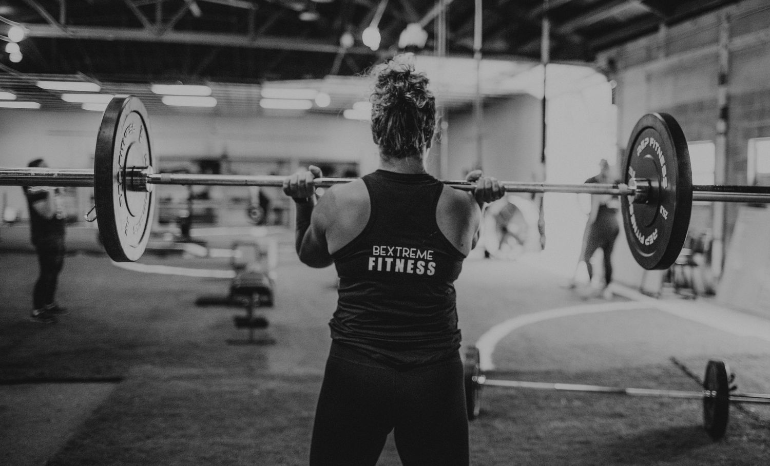 Athletes performing barbell lifts in a CrossFit gym with industrial lighting and equipment.