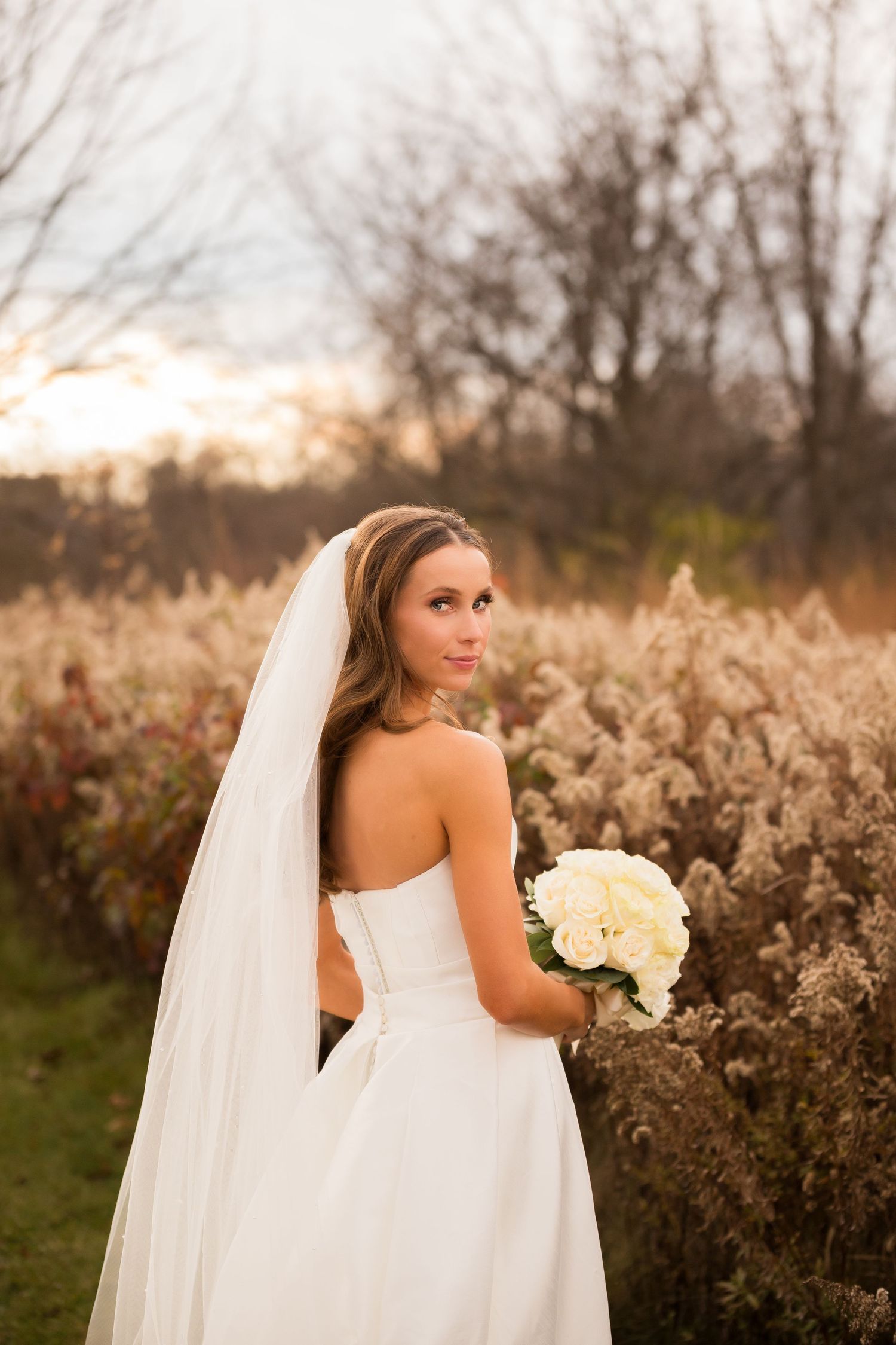 Bride holds white bouquet while posing in autumn field with long flowing veil.