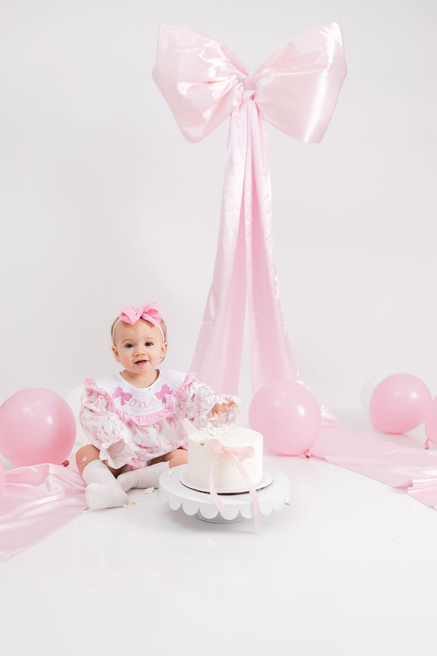 Baby in white dress surrounded by pink balloons and bows during milestone photo session.
