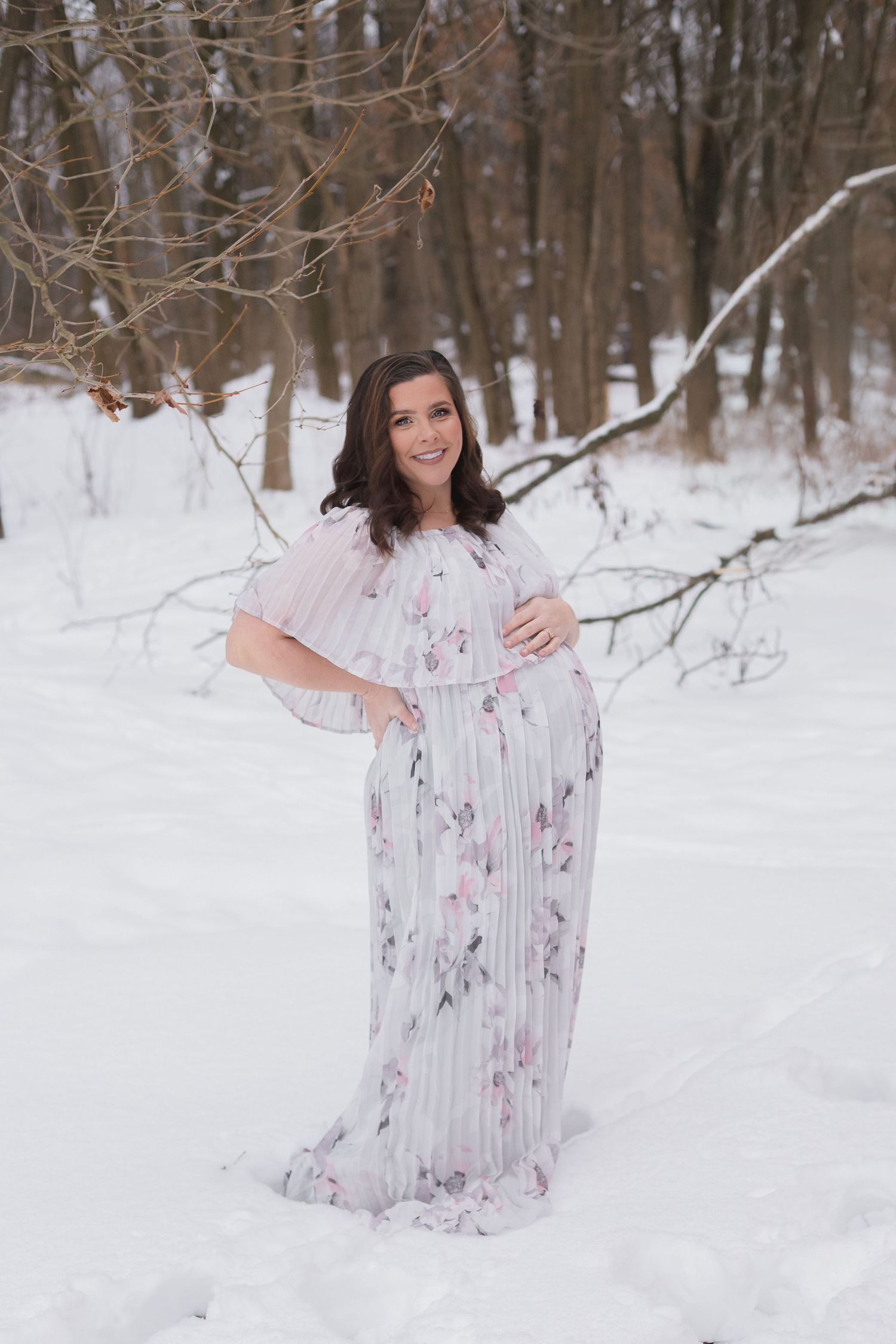 Maternity photos in snow covered woods with flowing floral dress.