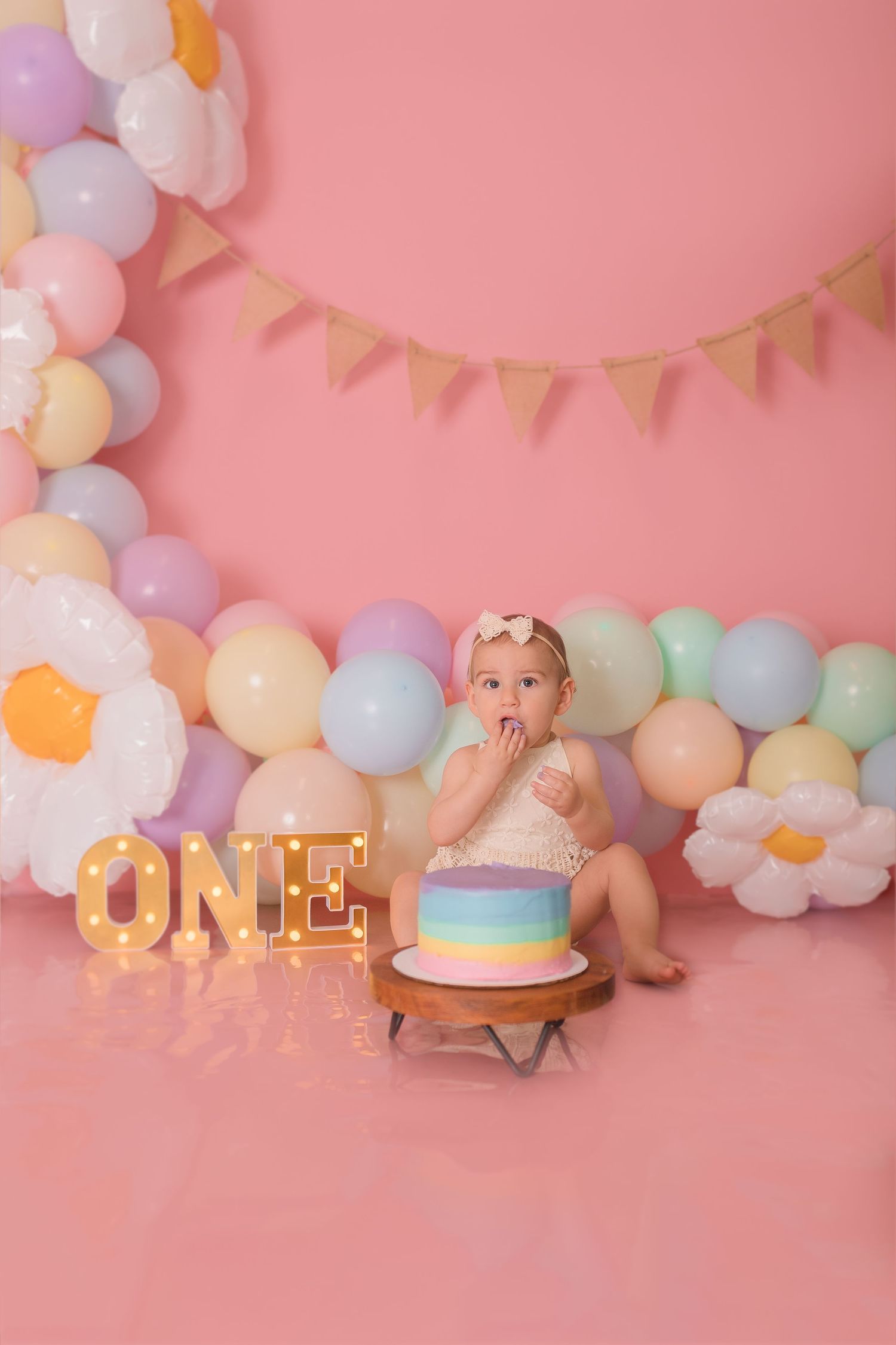 First birthday party setup with illuminated 'ONE' sign, rainbow cake, and pastel balloon decorations.