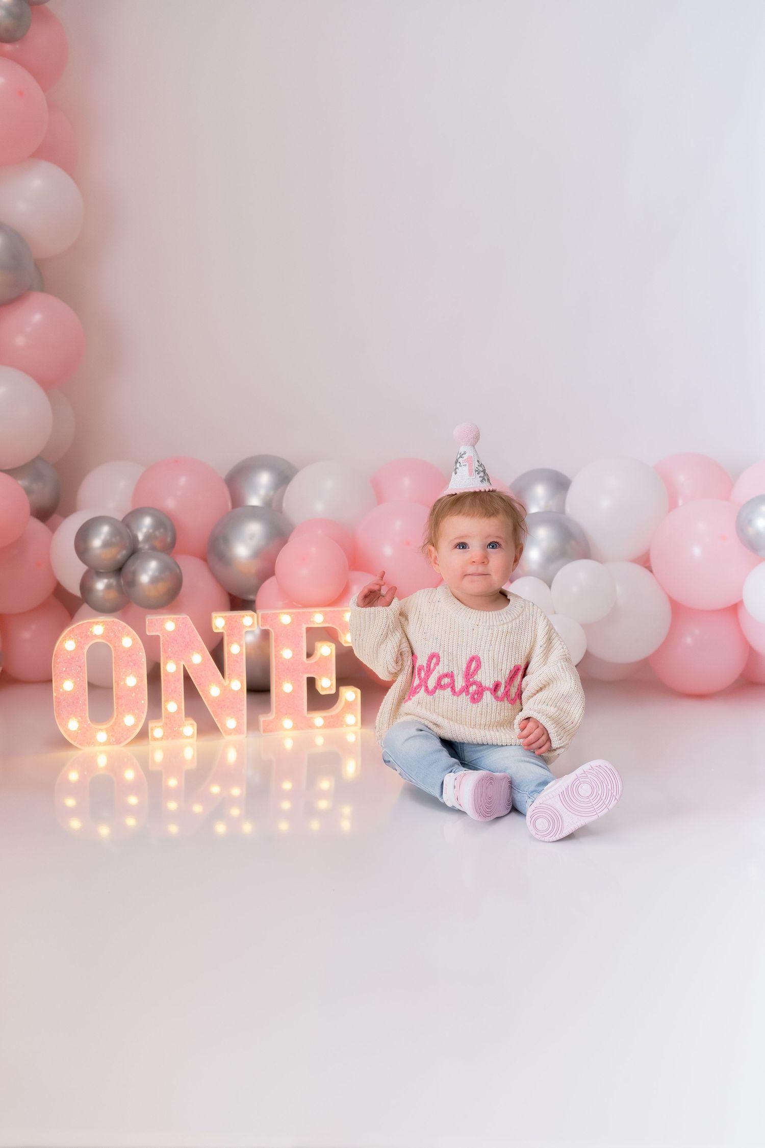 First birthday setup with illuminated ONE sign, balloons, and baby in cream sweater.