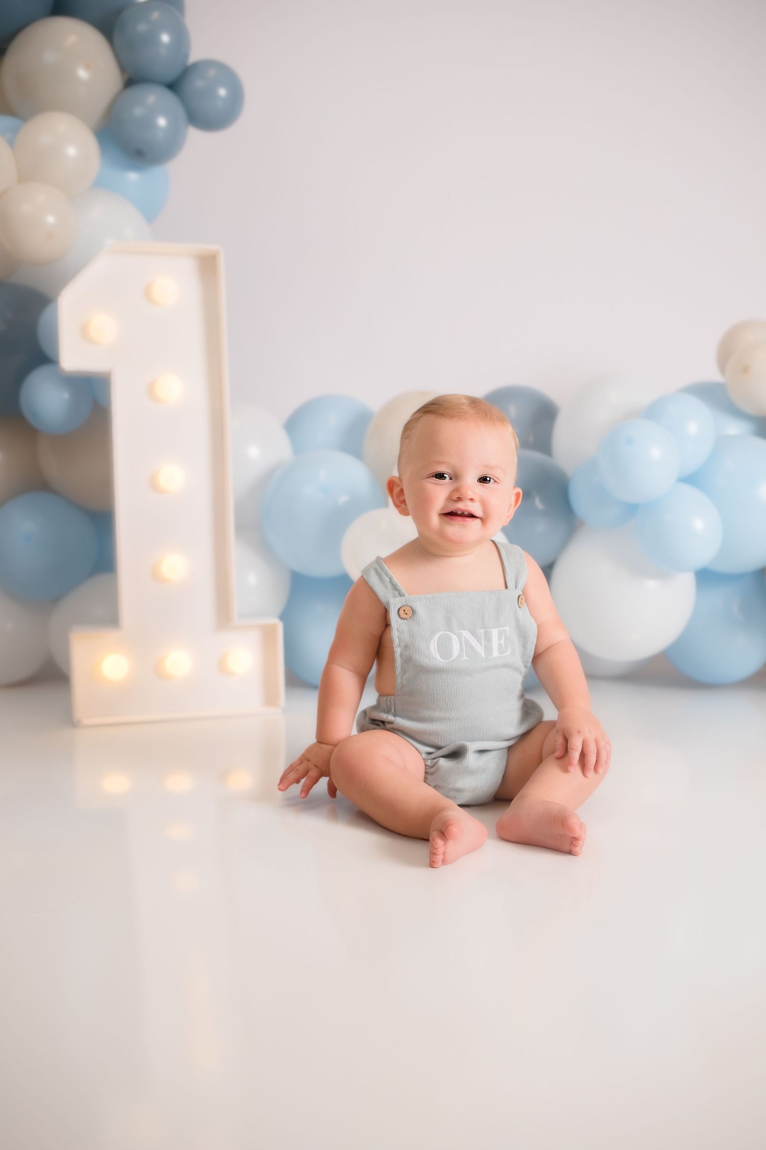 Baby in grey romper sitting by illuminated number one and blue balloons for first birthday photo.