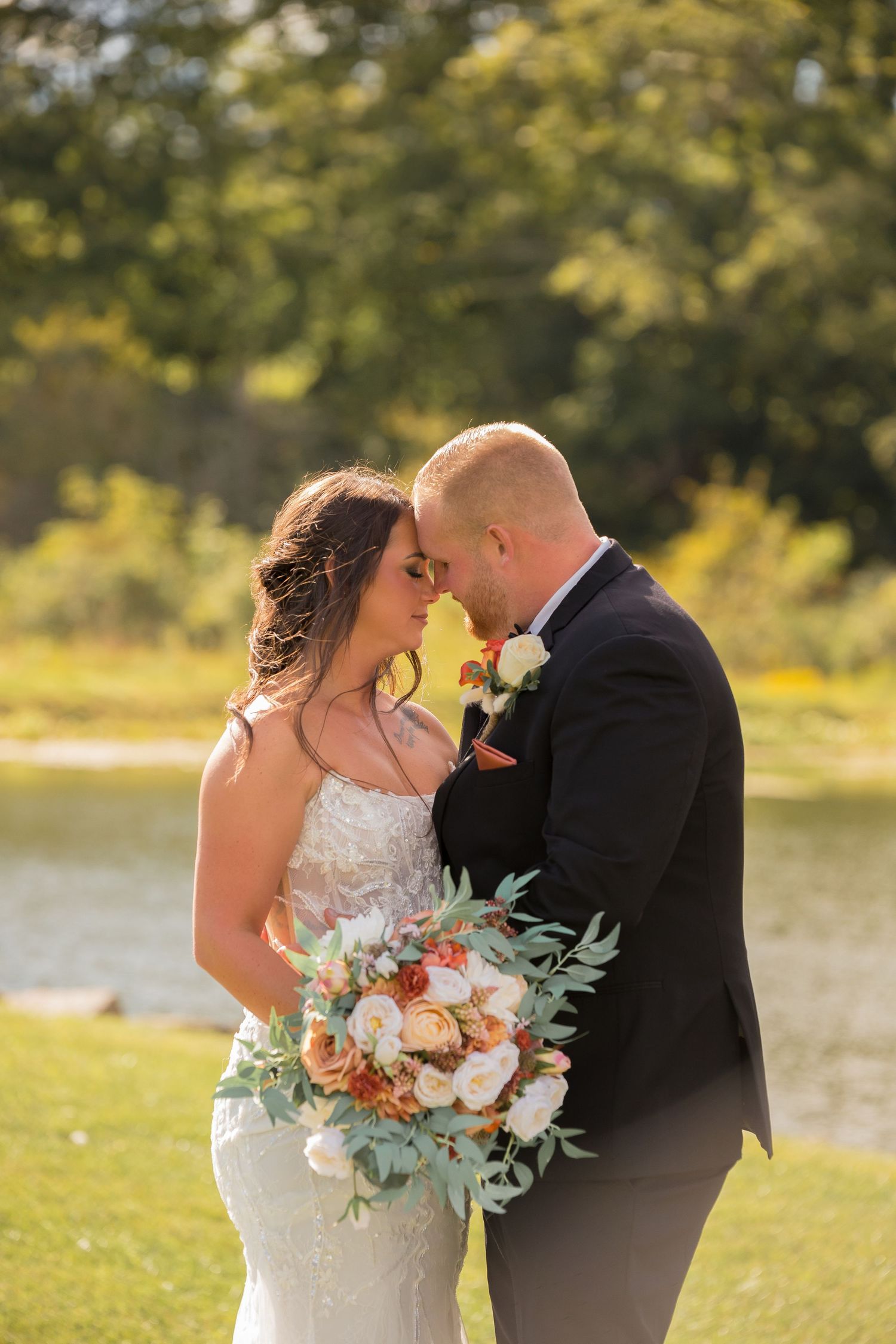 Romantic wedding couple shares intimate moments outdoors with a peach and white floral bouquet near a lake.
