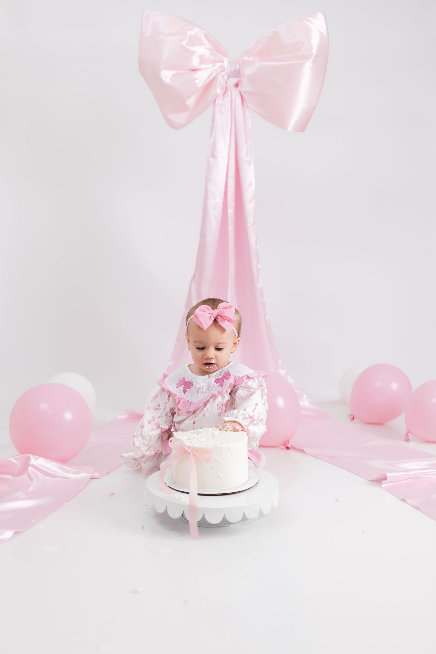 Pink first birthday scene with large bow backdrop, cake setup, and baby in floral outfit.