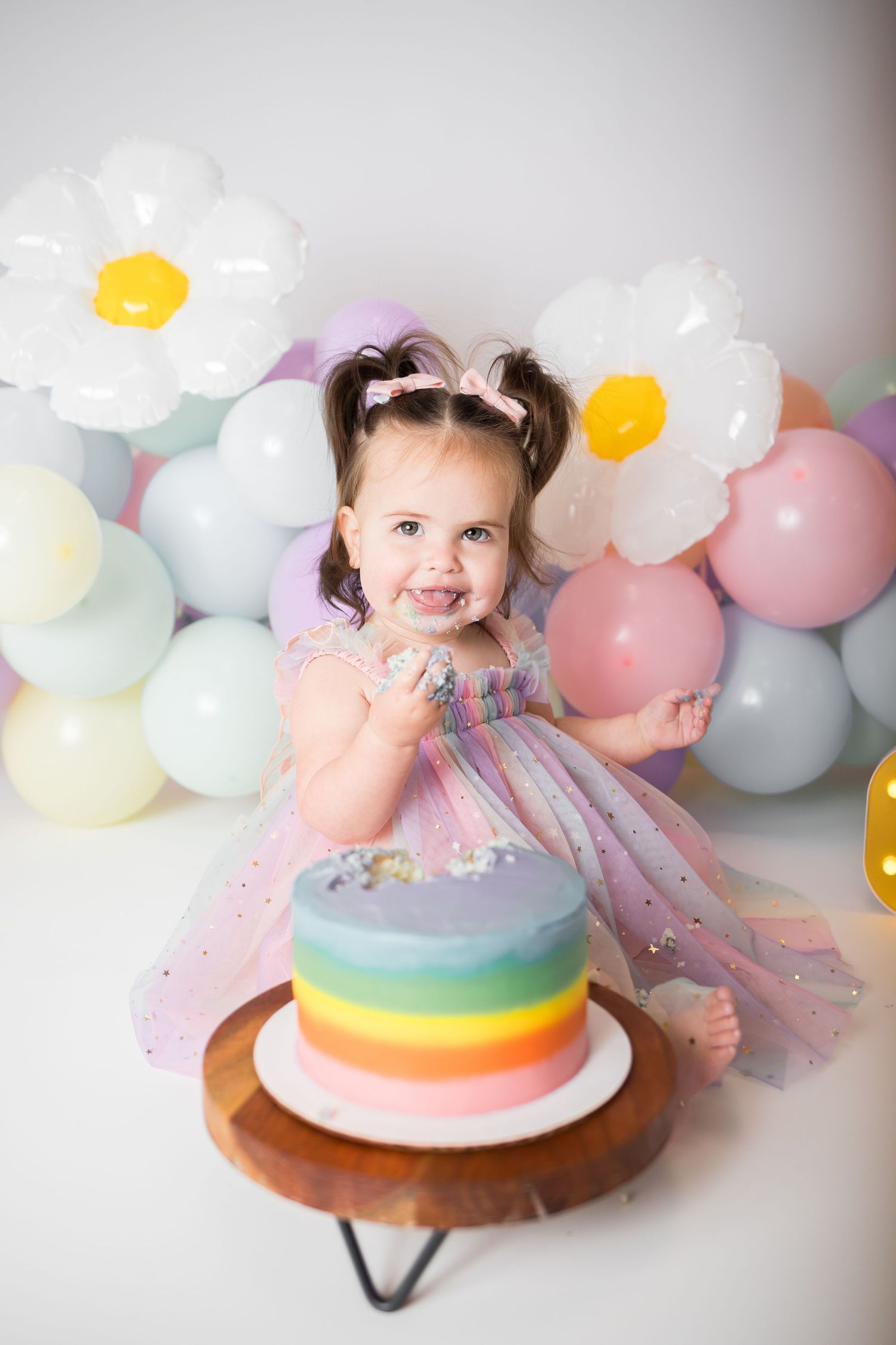 Festive scene with rainbow cake and baby in tulle dress amid pastel balloon decorations.