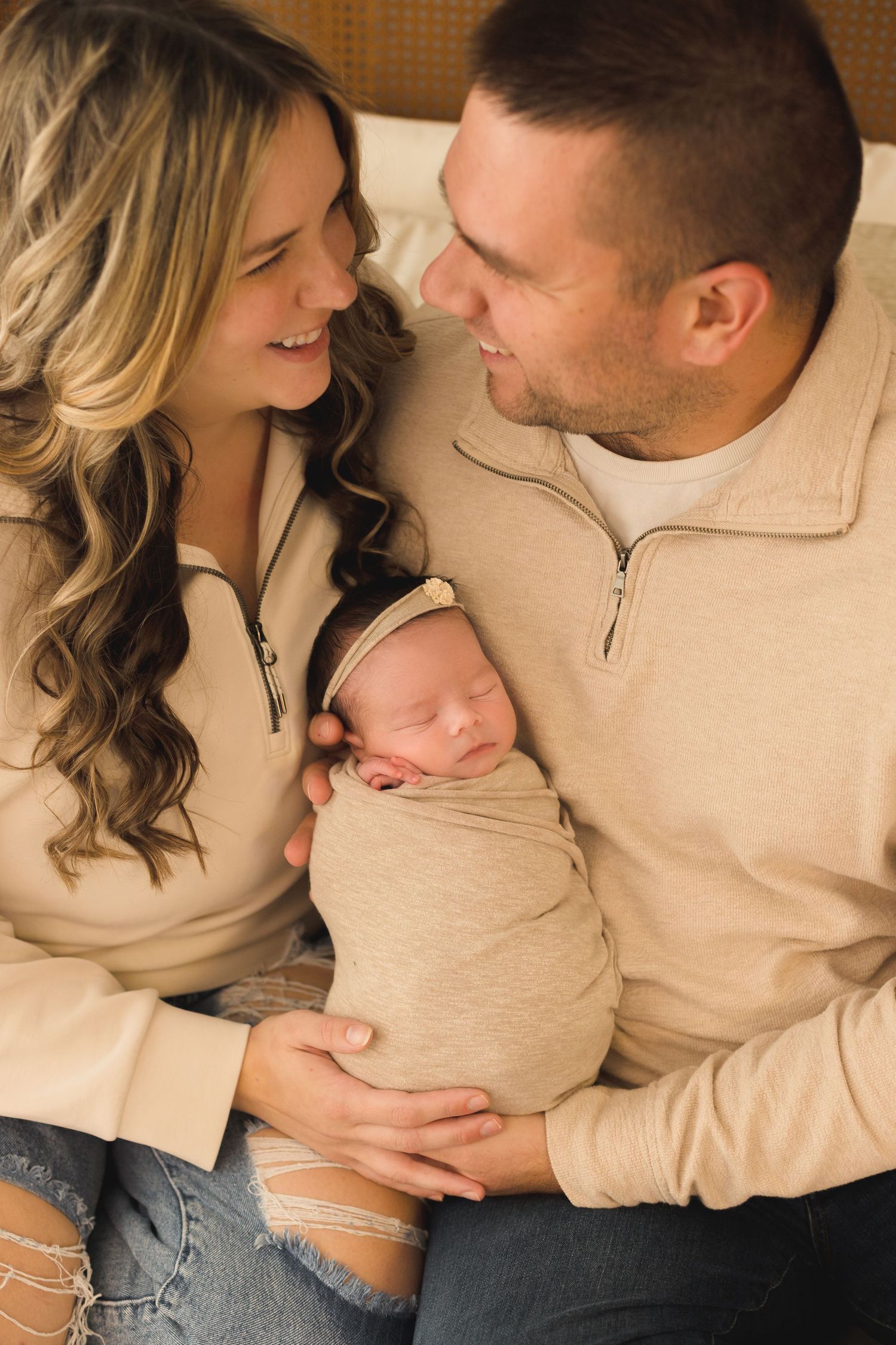 Parents in coordinating outfits share tender moment with newborn on cozy bed.