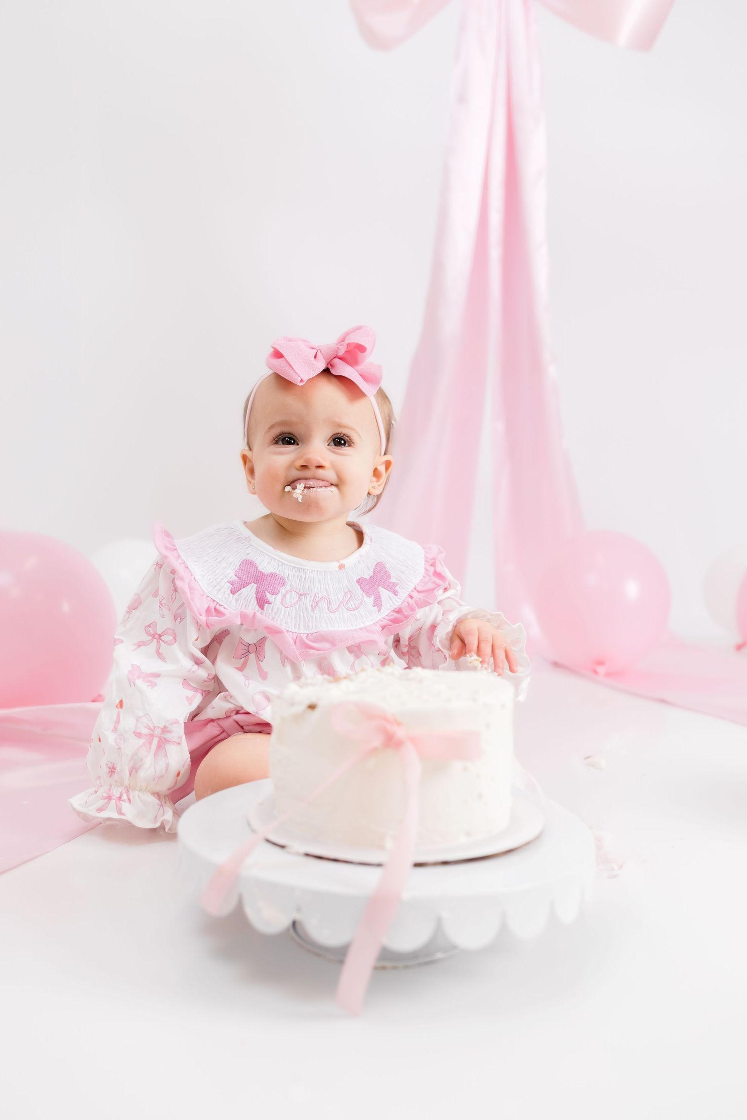 Baby in pink dress sitting on white floor with birthday cake during first birthday cake smash photo session.