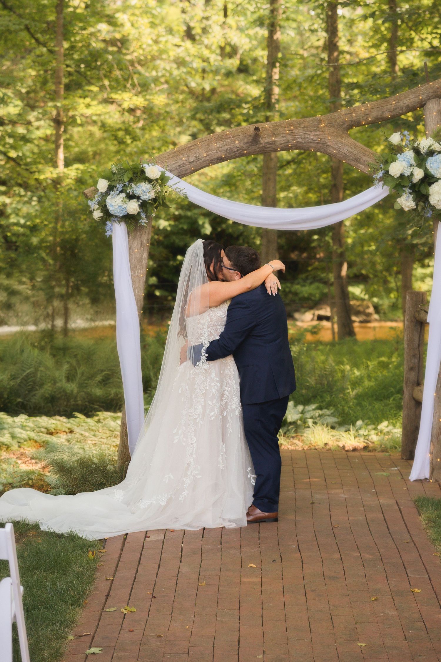 Wedding ceremony on brick pathway with white fabric draped arch and floral decorations.