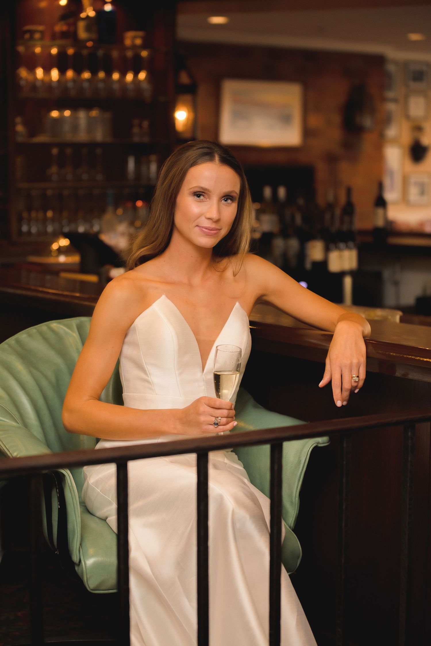 Bride in white gown holds champagne while seated at elegant bar.