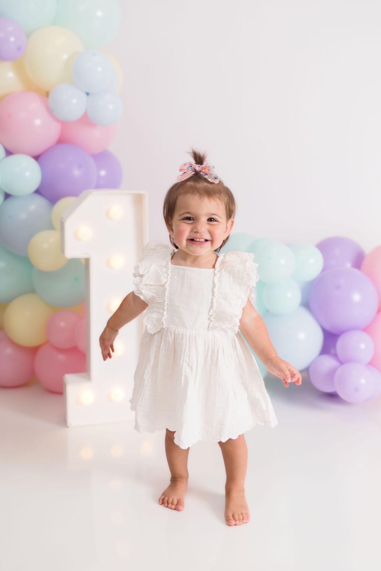 Baby in white dress dancing among pastel balloons at first birthday celebration.