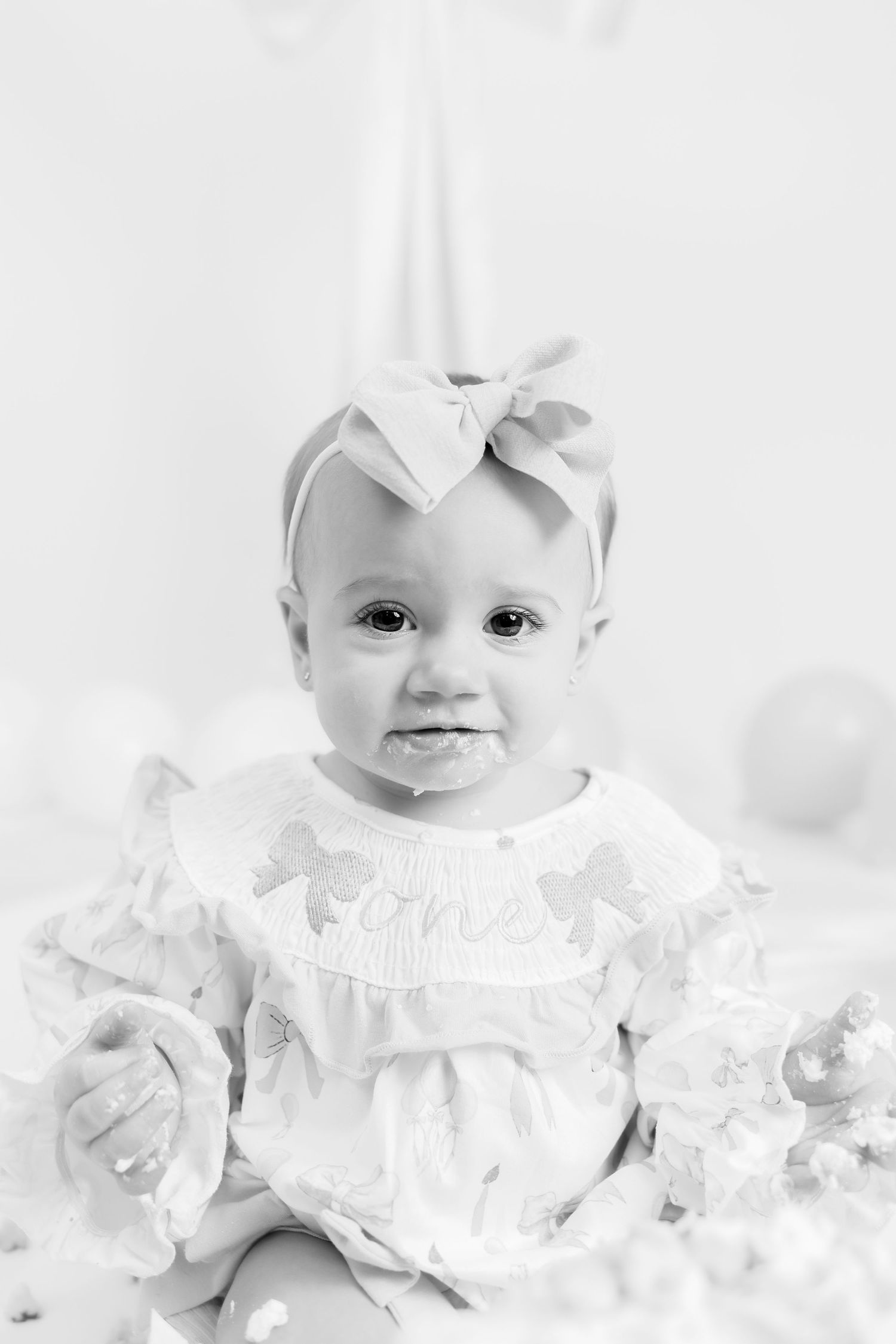 Baby in white ruffled dress and bow poses against white background in classic portrait style.