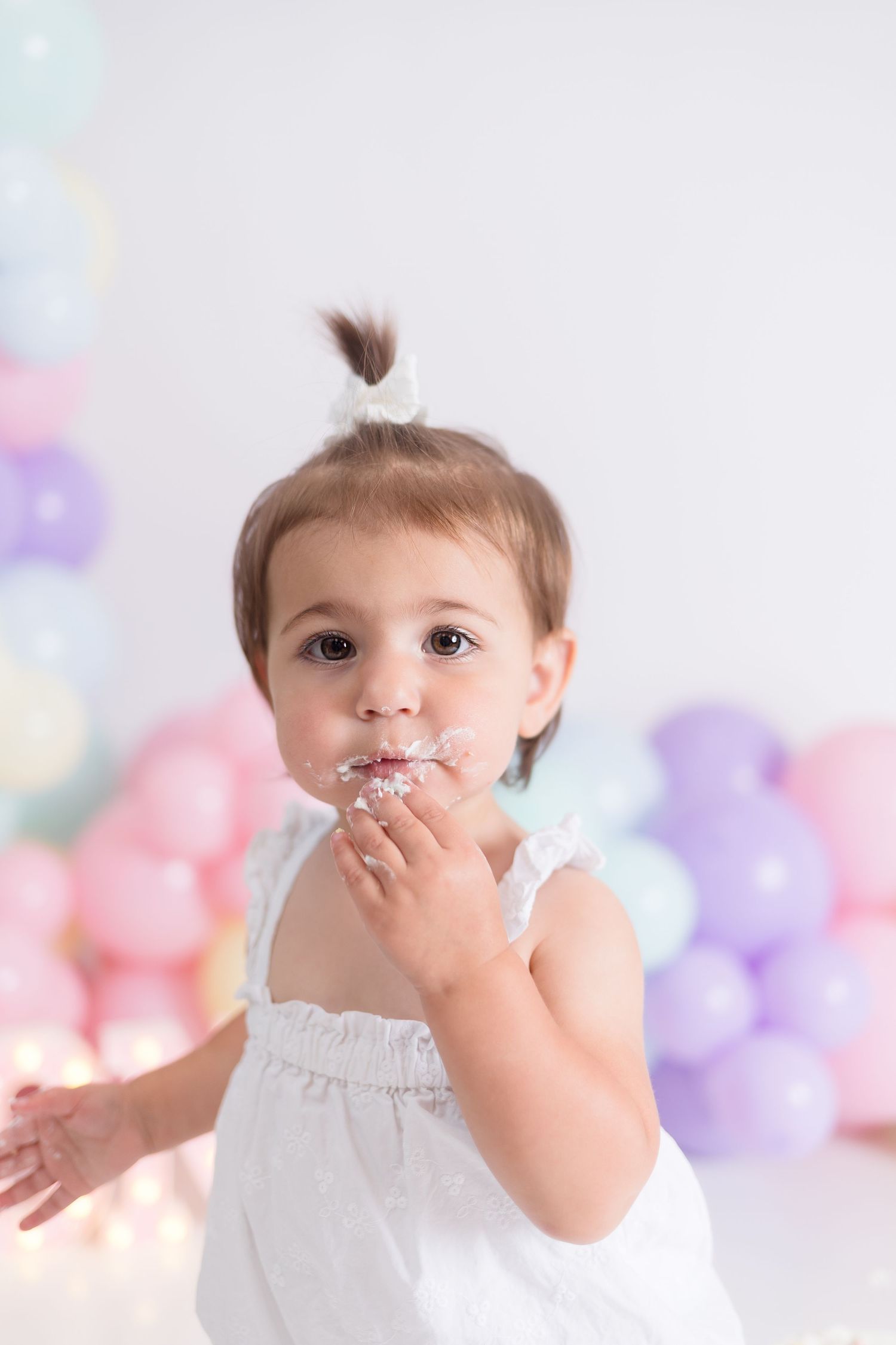 Baby in white dress with pastel balloon backdrop enjoying cake.