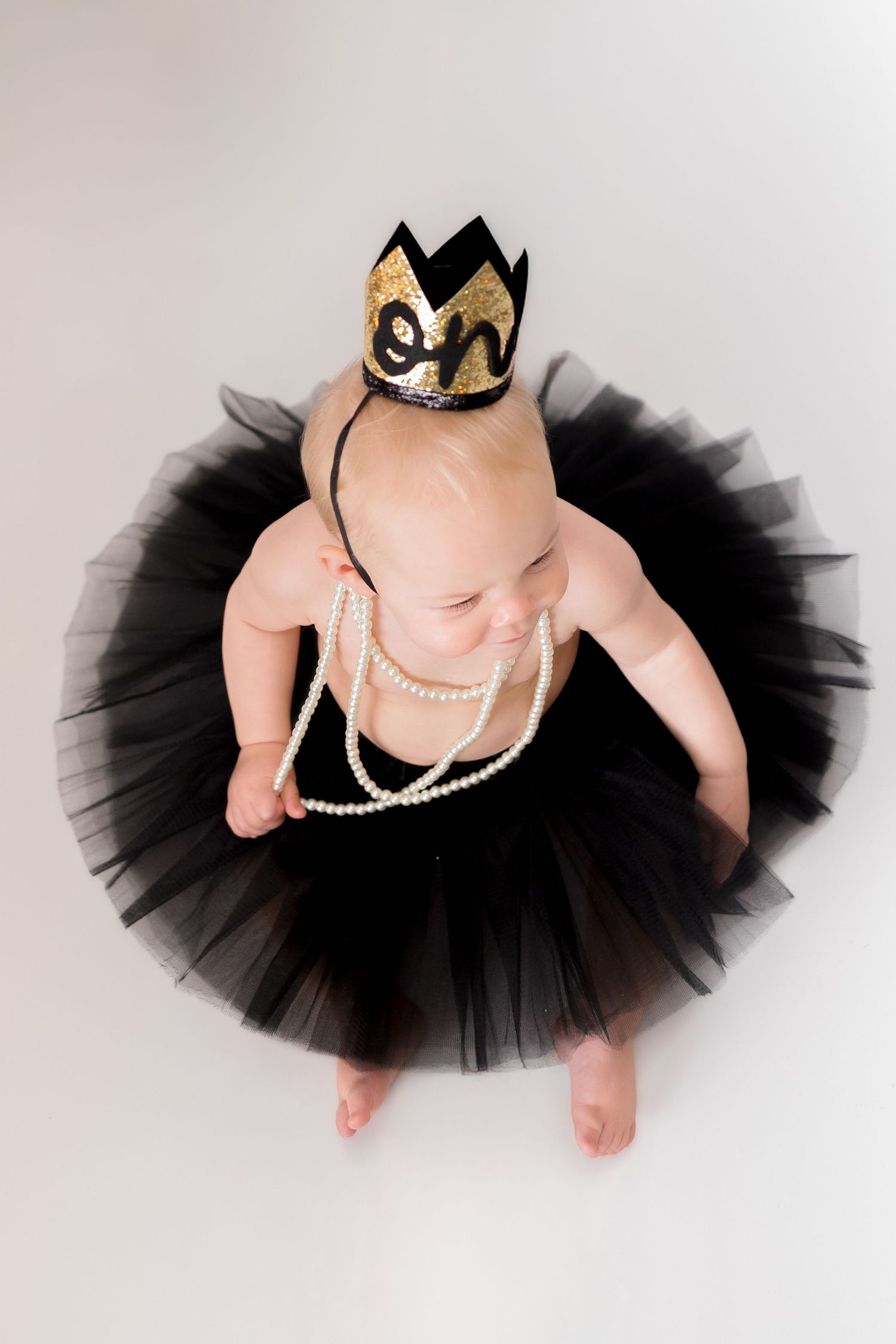 Overhead view of baby wearing black tutu and gold crown with pearl necklaces.
