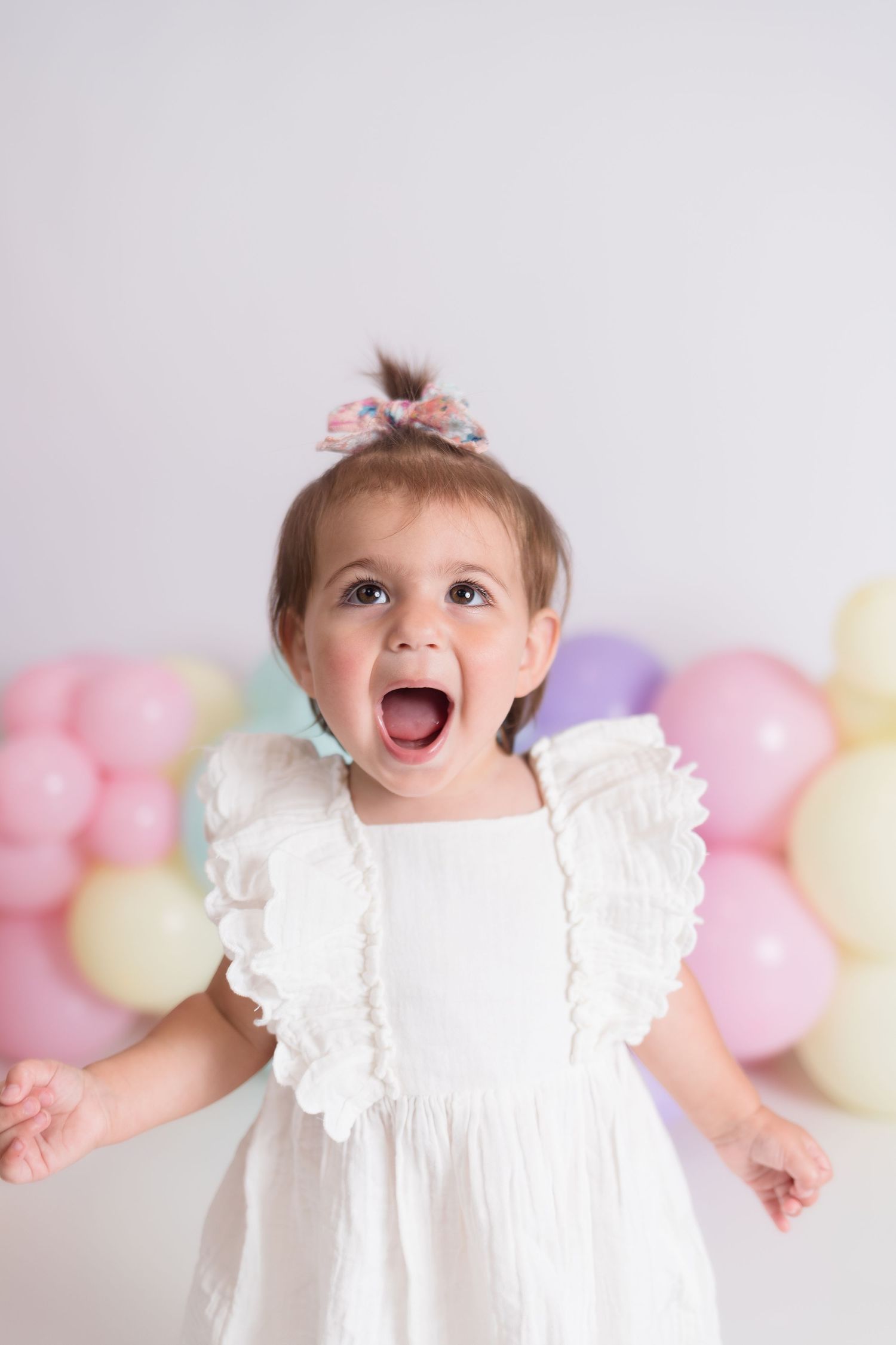 Baby in white lace dress smiling against pastel balloon background.