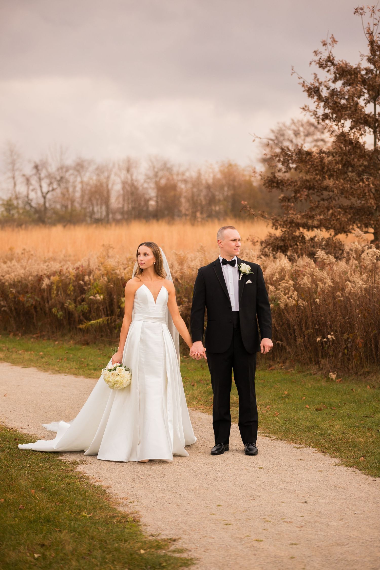 Newlyweds walk hand in hand on path surrounded by golden autumn grasses.
