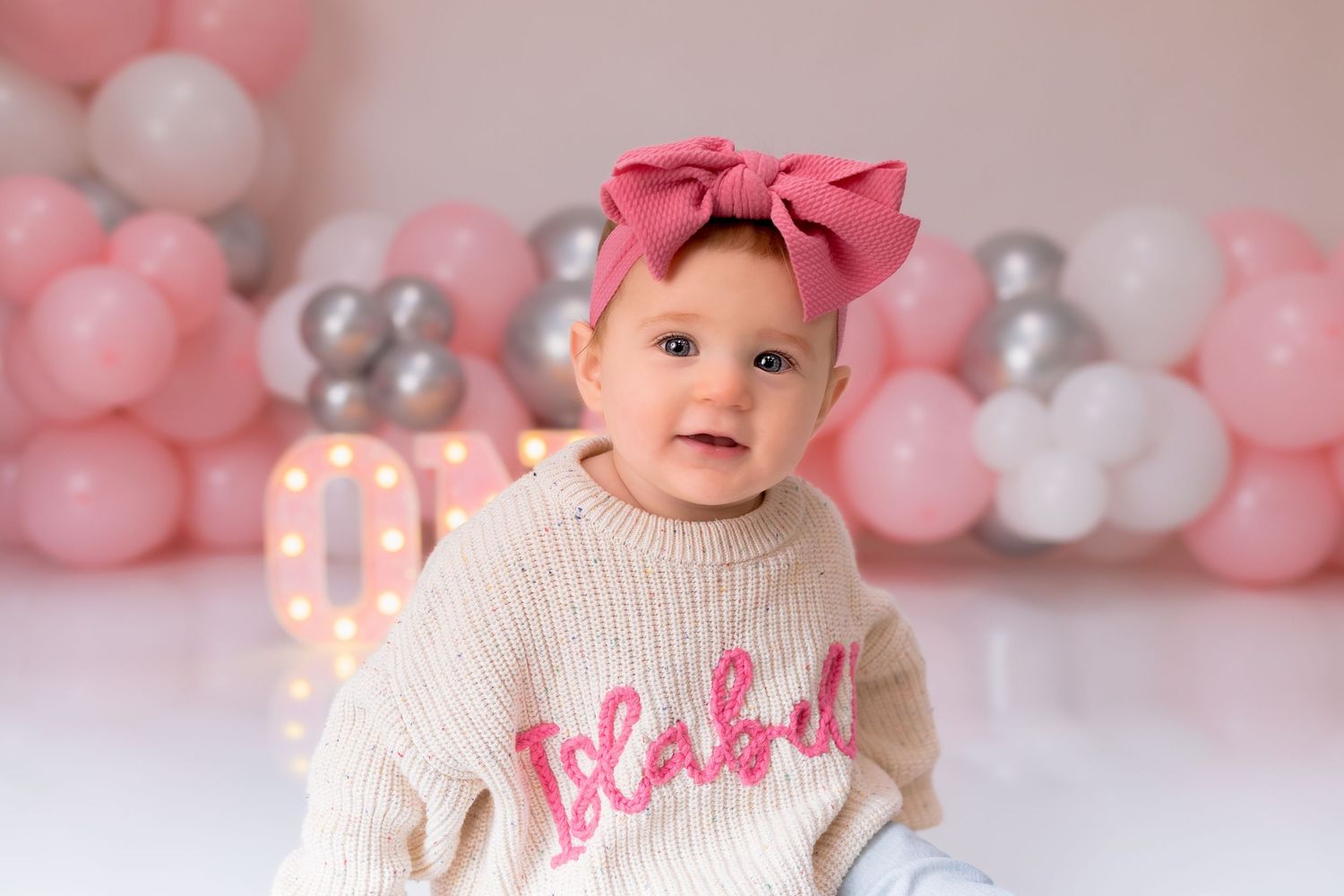 Baby in cream sweater with pink text and bow posing with pink and silver balloons.