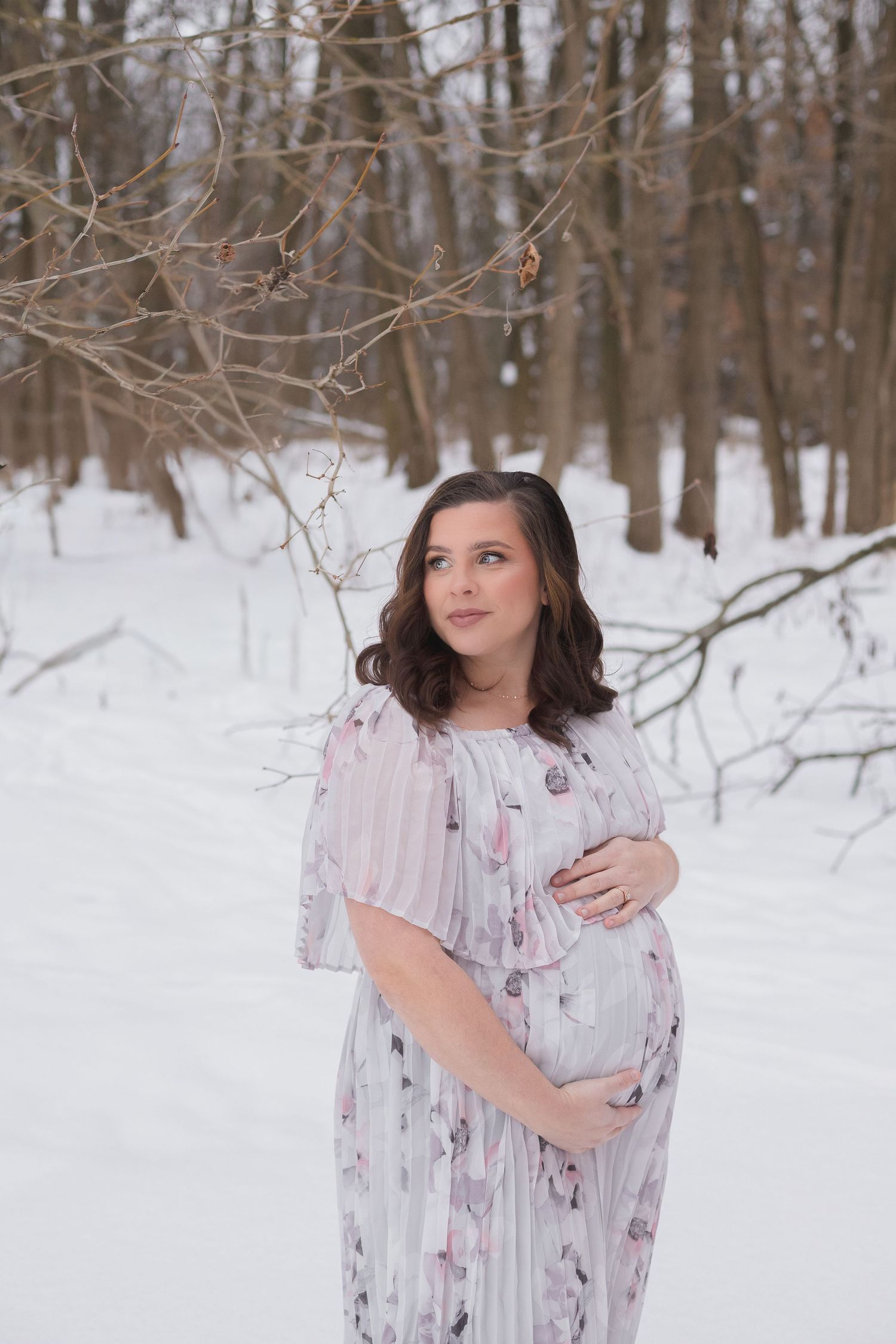 Winter maternity portrait in snowy forest wearing floral white dress.
