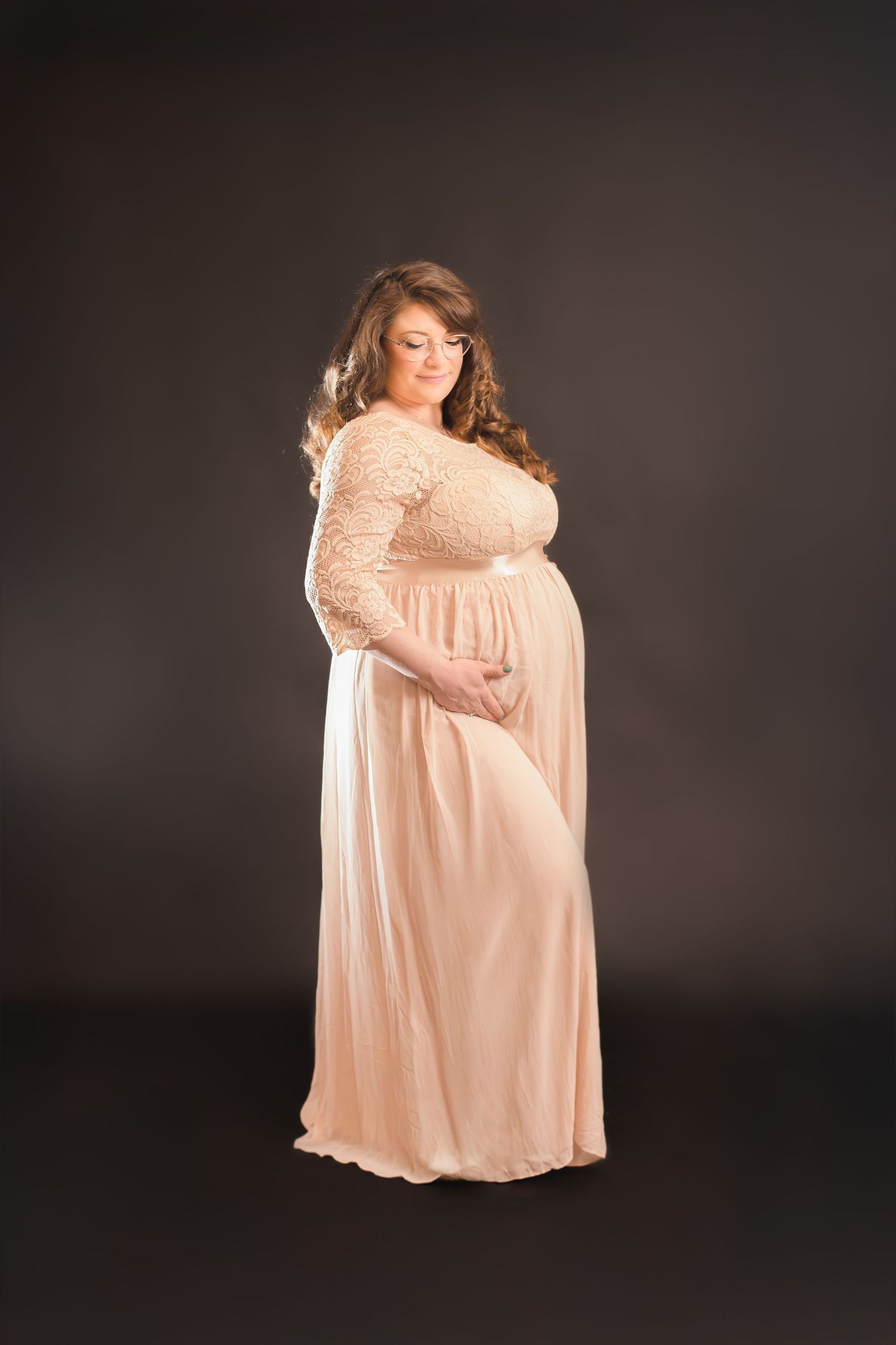 Maternity photoshoot in peach dress against dark background showing pregnancy silhouette.