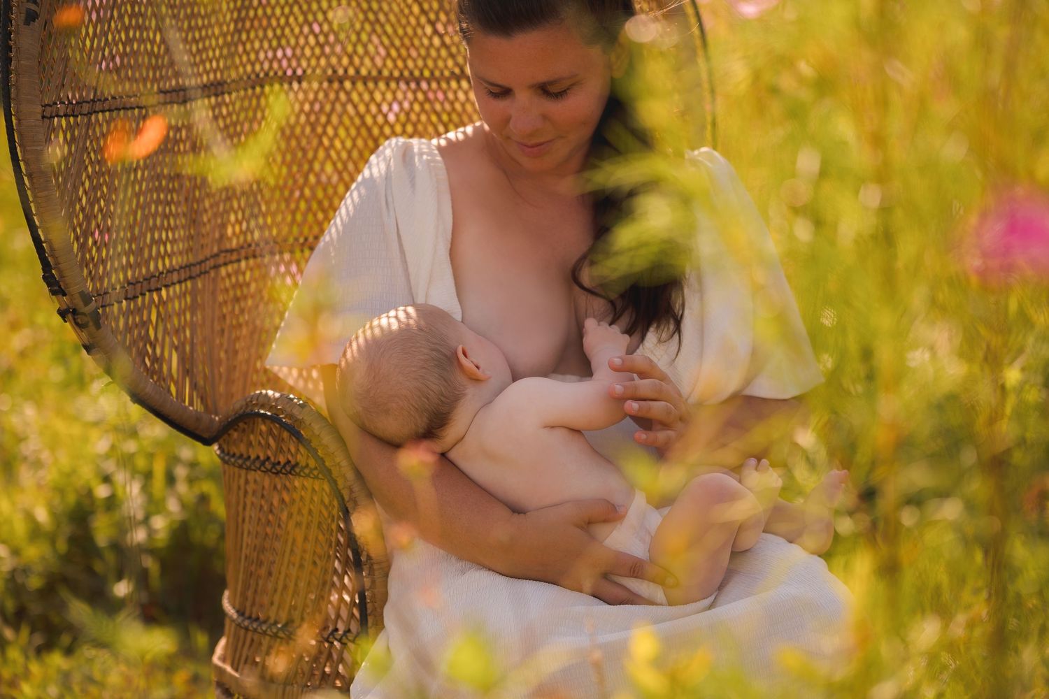 Intimate nursing portrait in a sunlit wildflower field with a vintage peacock chair.