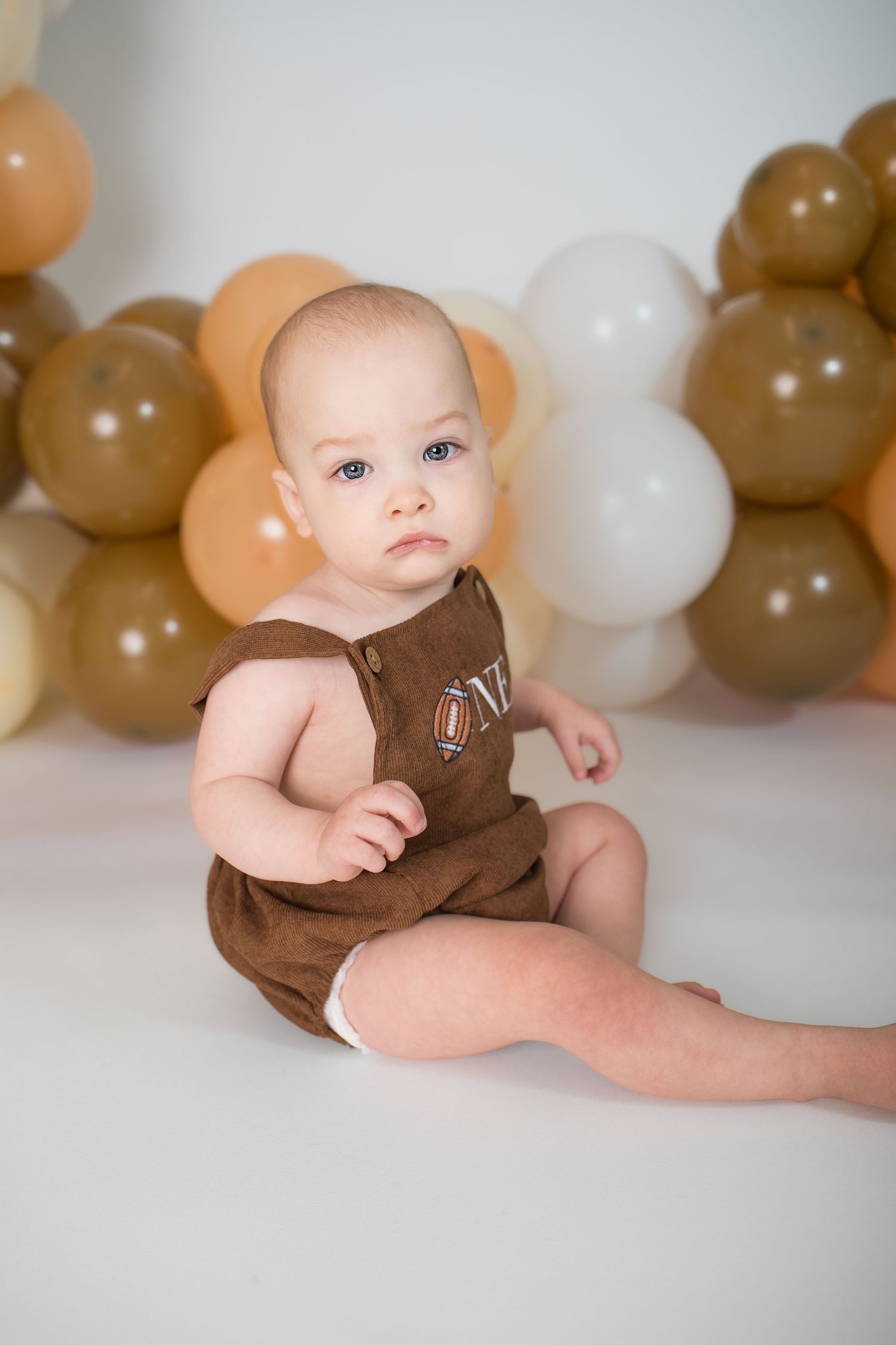 Baby in brown outfit sitting against neutral balloon backdrop for milestone photo.