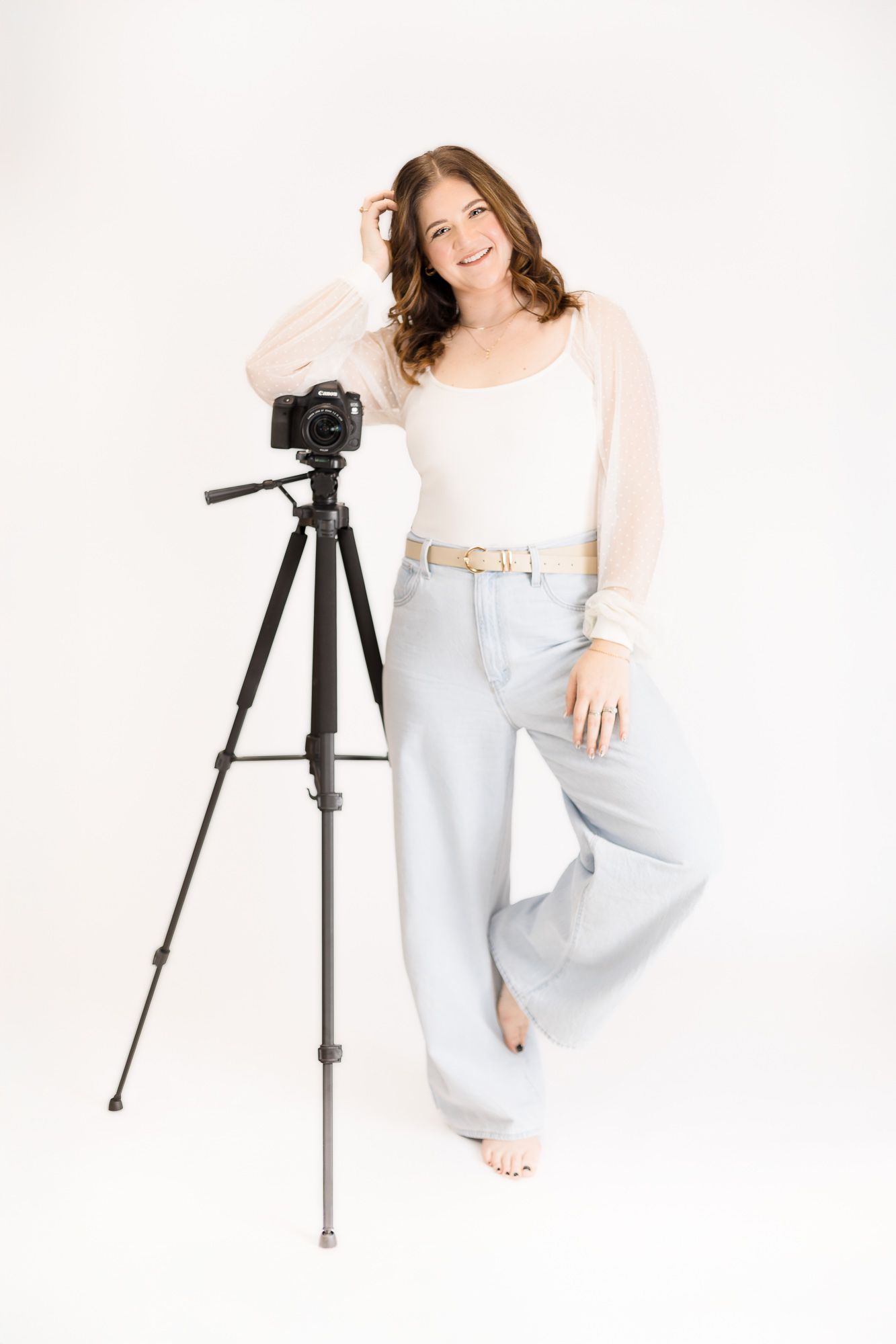 Woman in white top poses casually against white backdrop with camera tripod in studio setting.