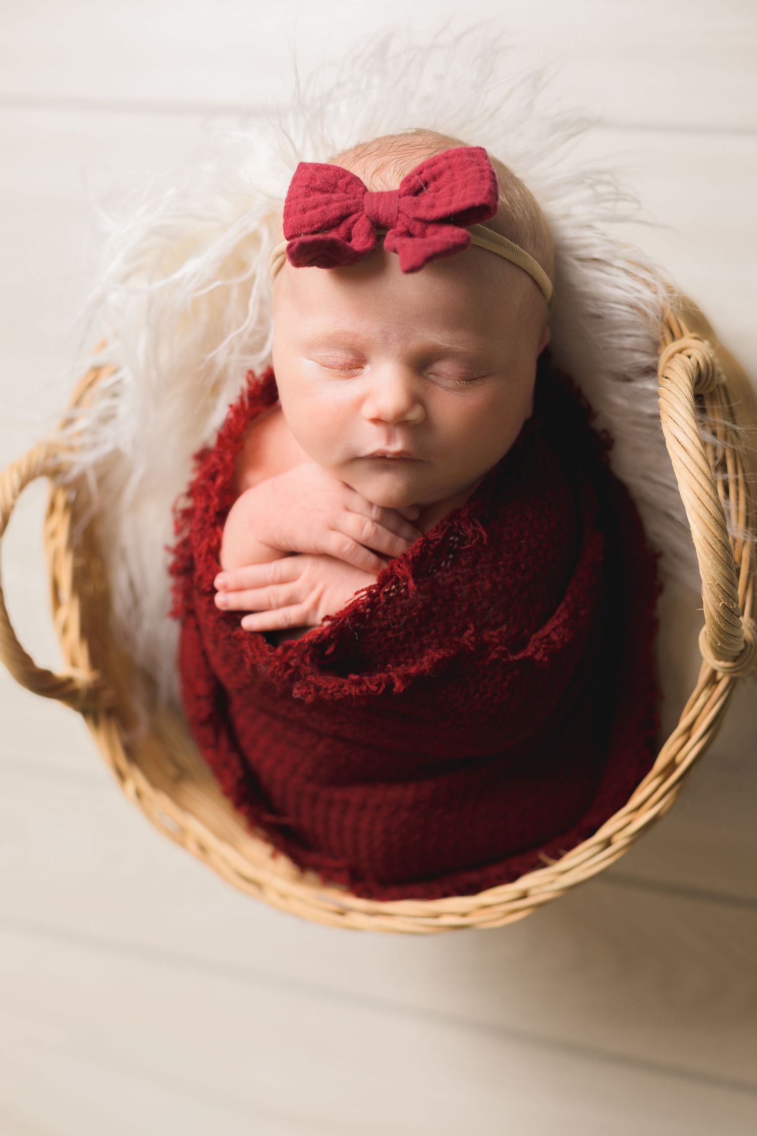 Sleeping newborn baby girl swaddled in red wrap with bow headband, nestled in a wicker basket with white fur.