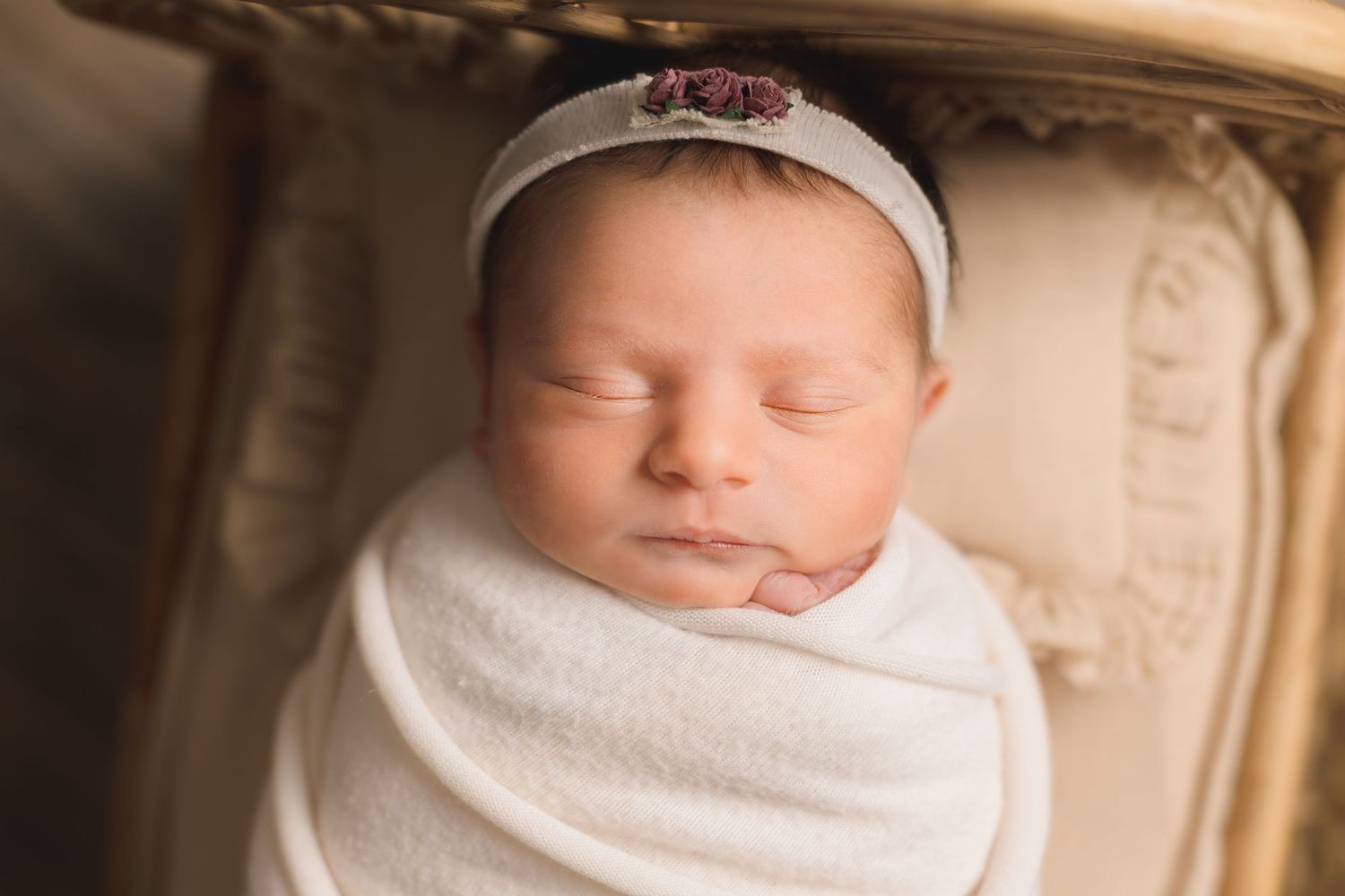A sleeping newborn baby wrapped in white swaddling cloth lies peacefully against a cream-colored background.