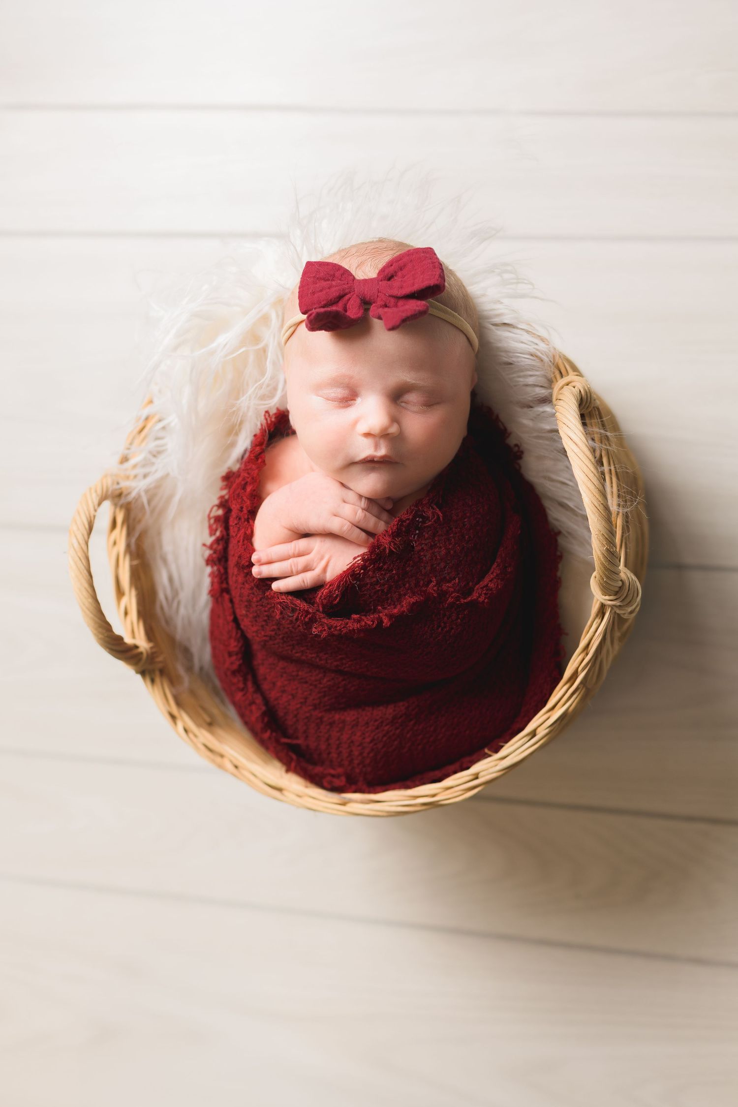 Newborn baby girl sleeping in a wicker basket, wrapped in red knit blanket with matching bow headband.