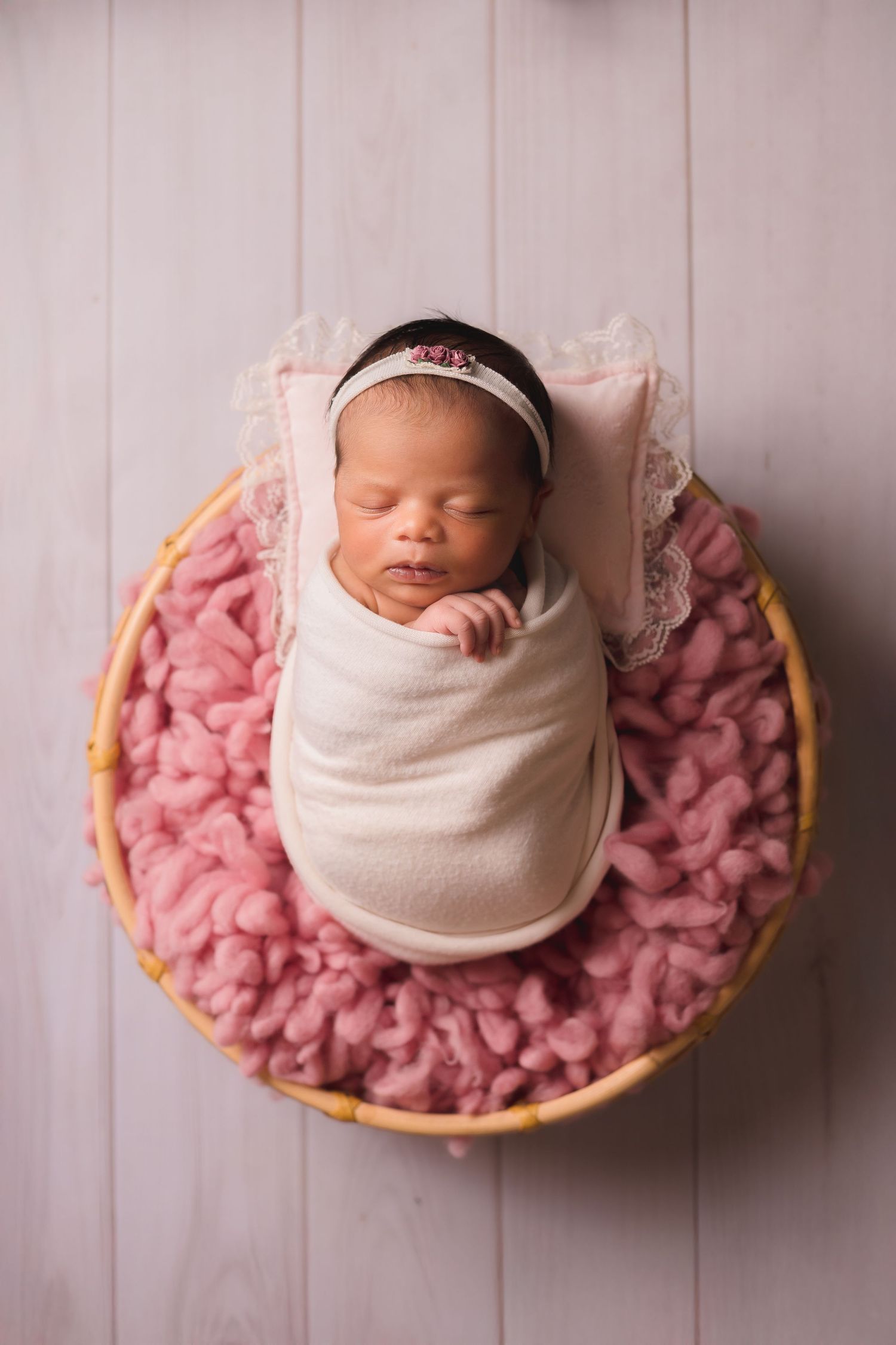 Newborn sleeping in basket with pink wool backdrop and cream wrap.
