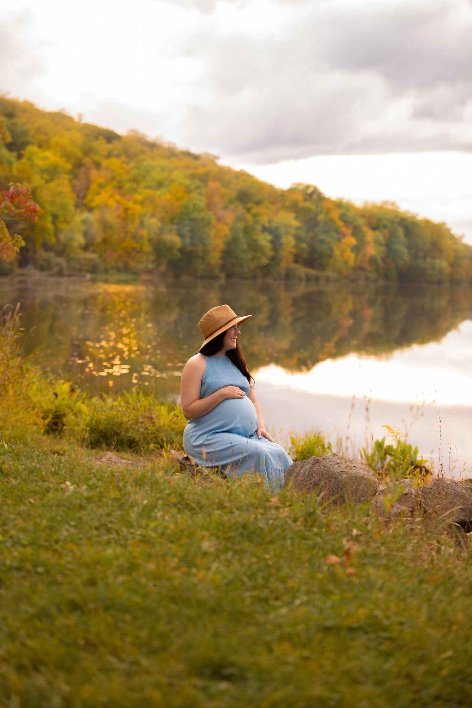 Person in blue dress and hat sits peacefully by lake surrounded by autumn colors.