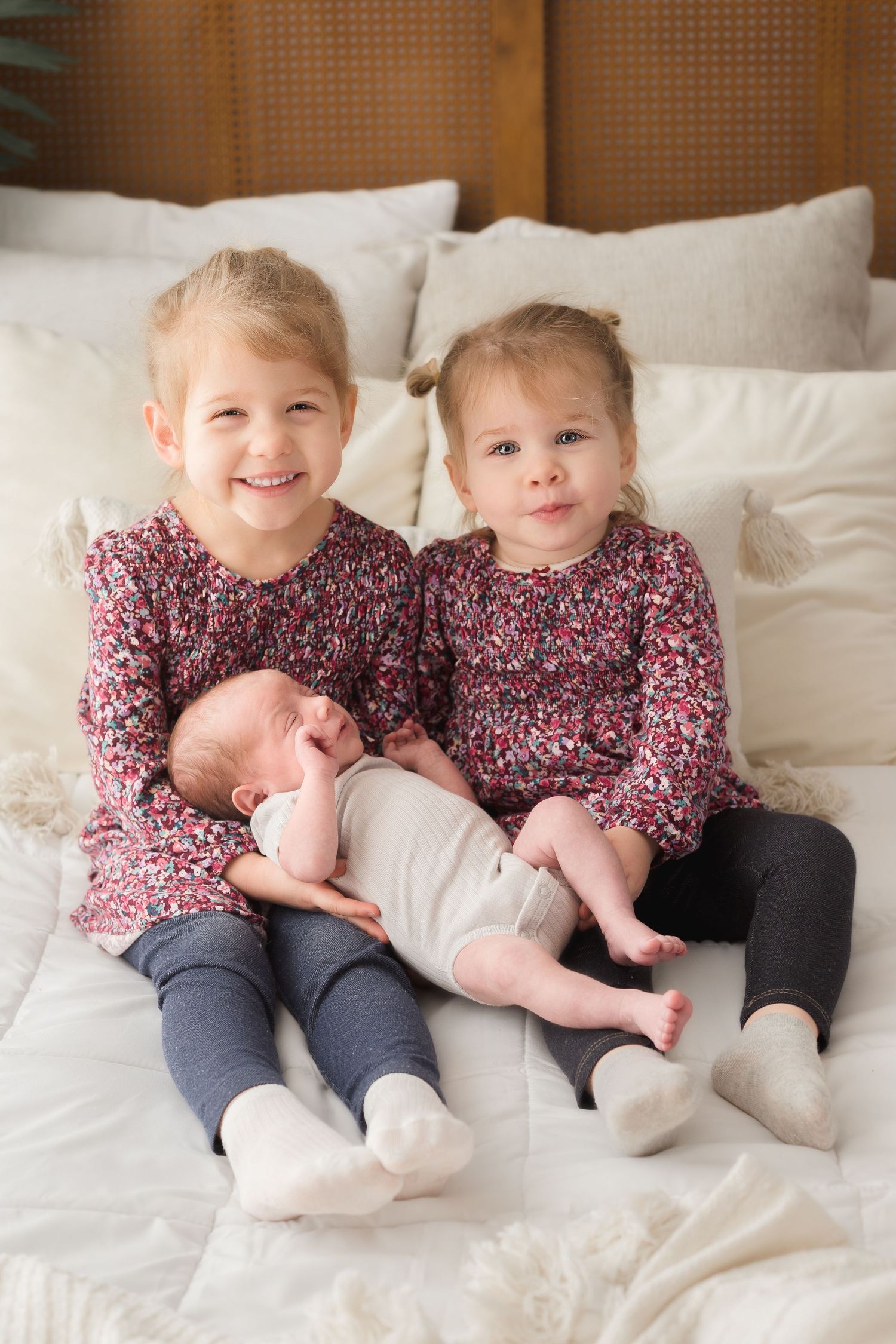 Siblings in matching floral dresses sitting together on white bed.