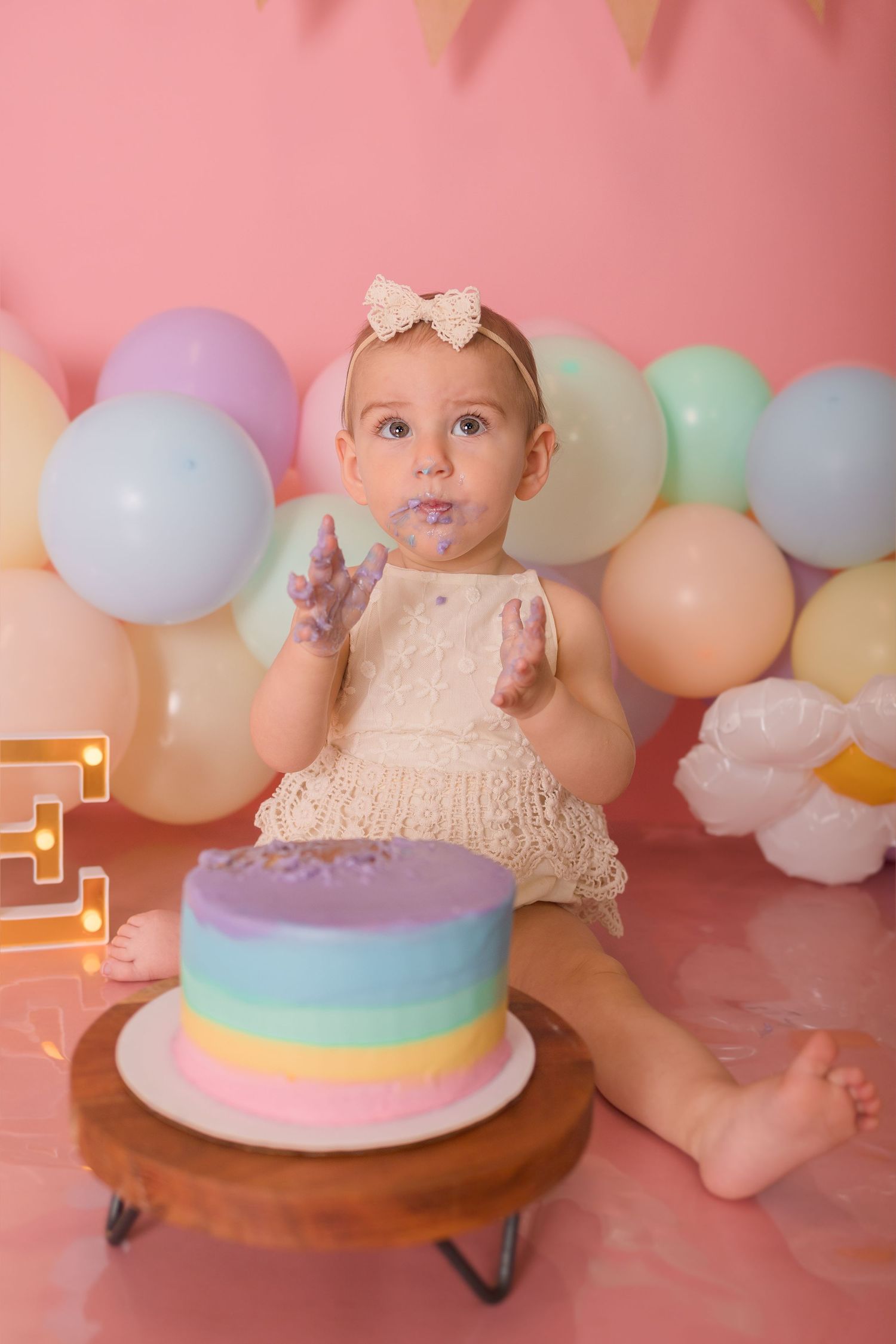 A festive first birthday setup with a rainbow cake and pastel balloon decorations on pink background.