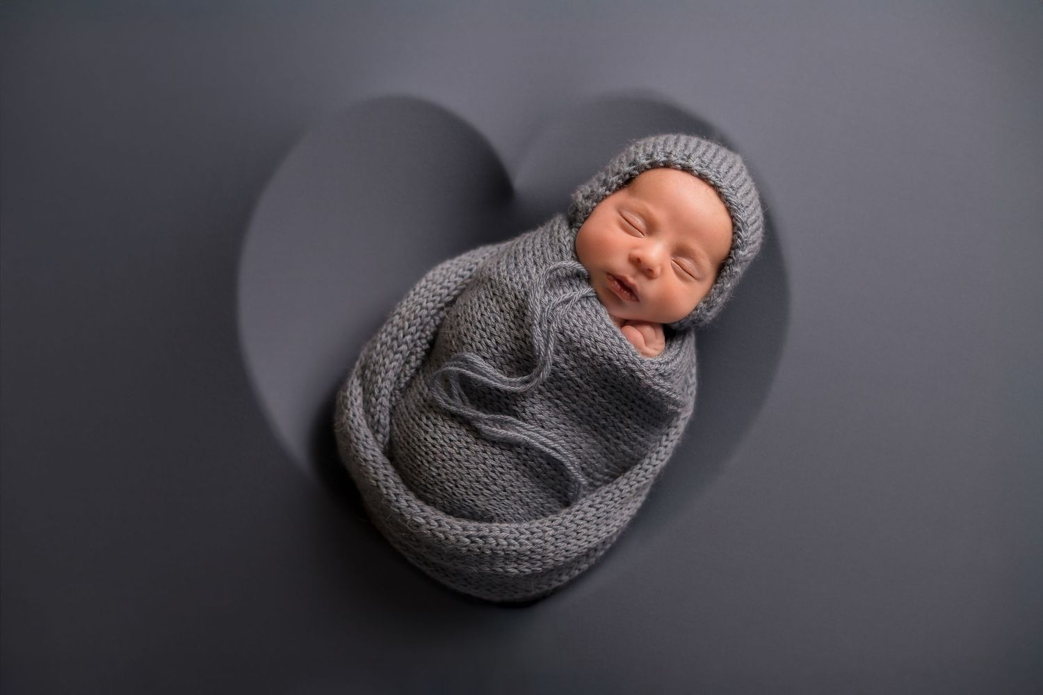 Sleeping infant wrapped in grey knit blanket against dark background.