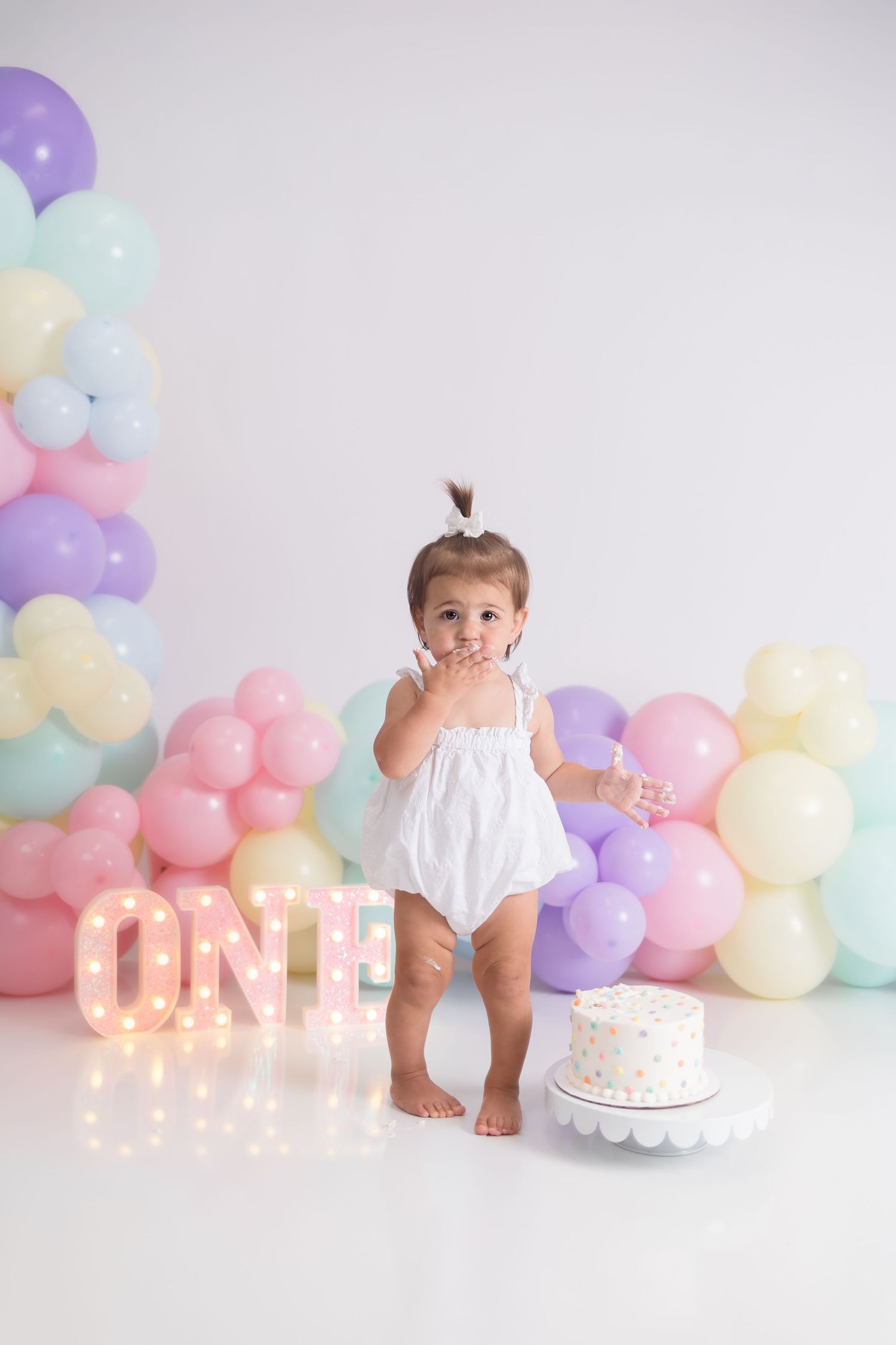 First birthday setup with illuminated letters, cake, and pastel balloon arch.