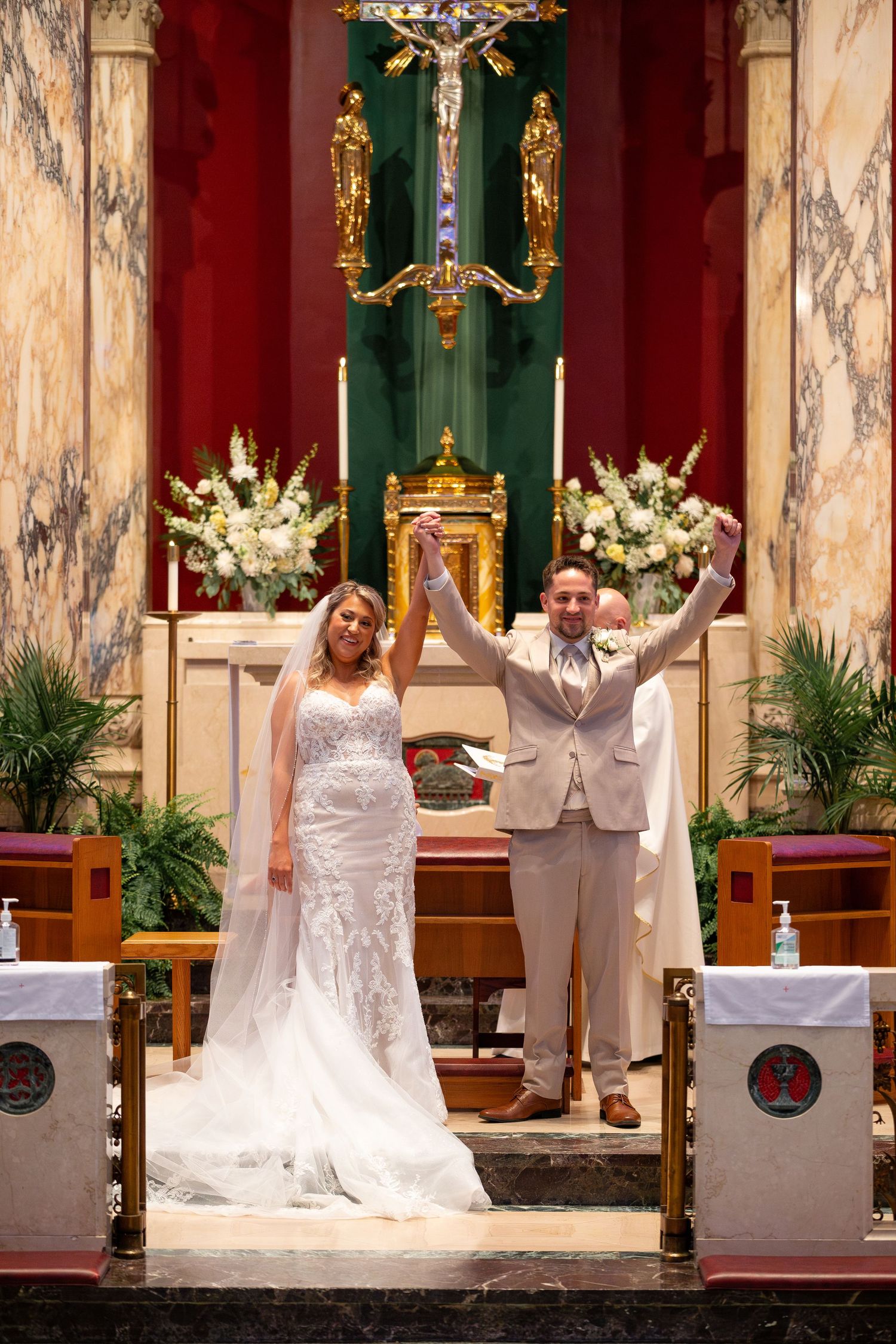 A wedding ceremony in an ornate church with marble columns and floral arrangements at the altar.