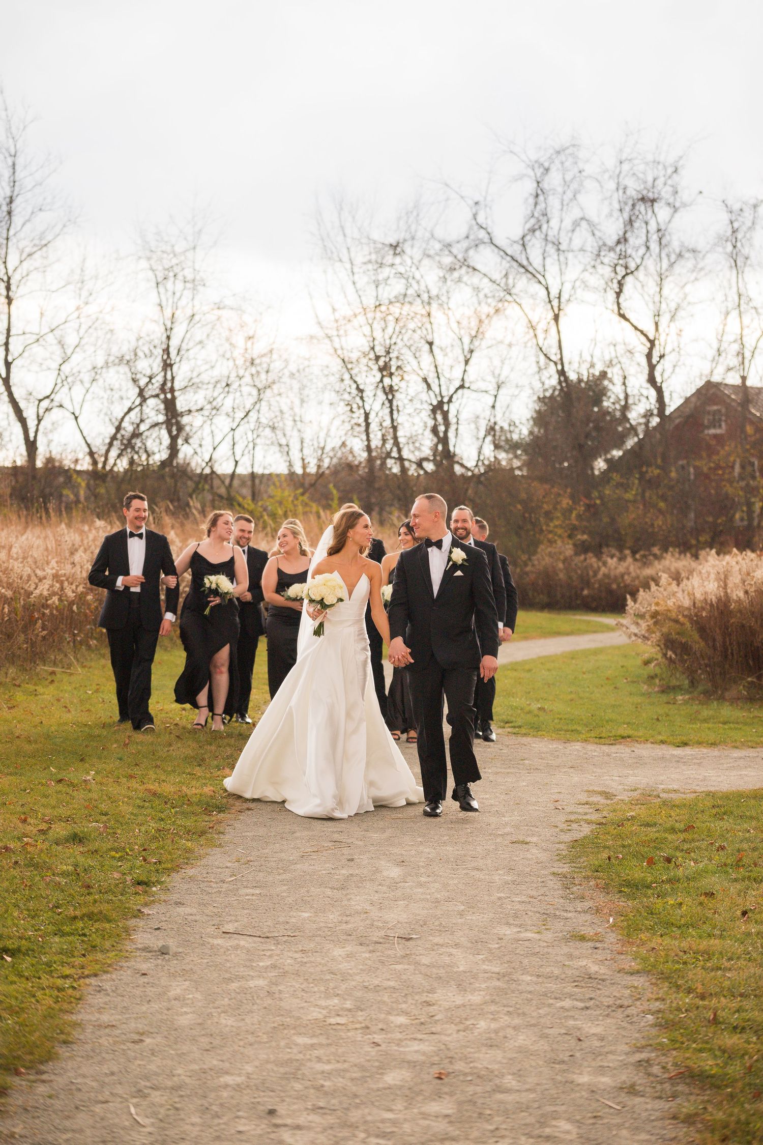 Wedding party walks along outdoor path during autumn with bare trees in background.