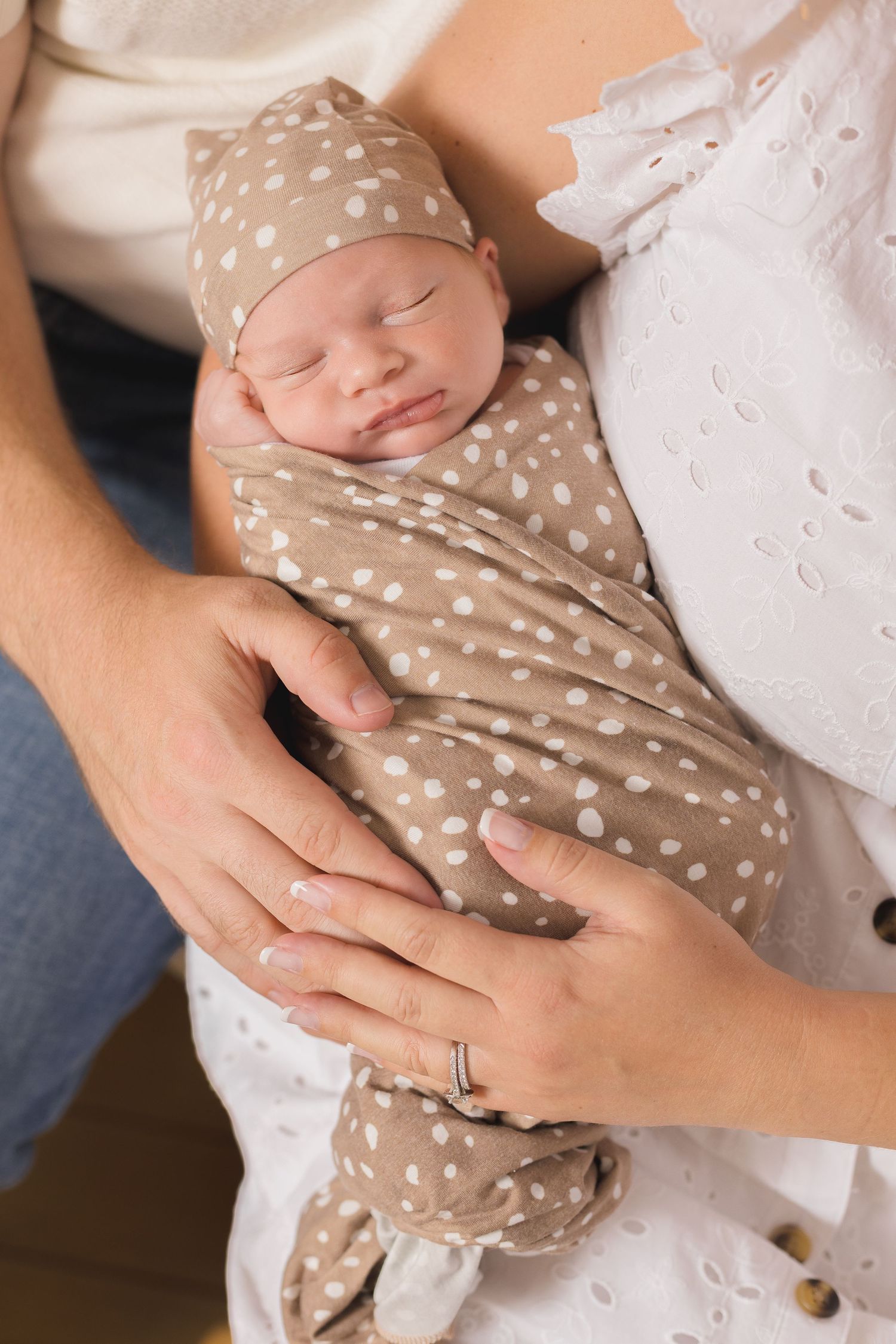 Newborn baby swaddled in tan polka dot blanket and hat, sleeping peacefully in parents arms.