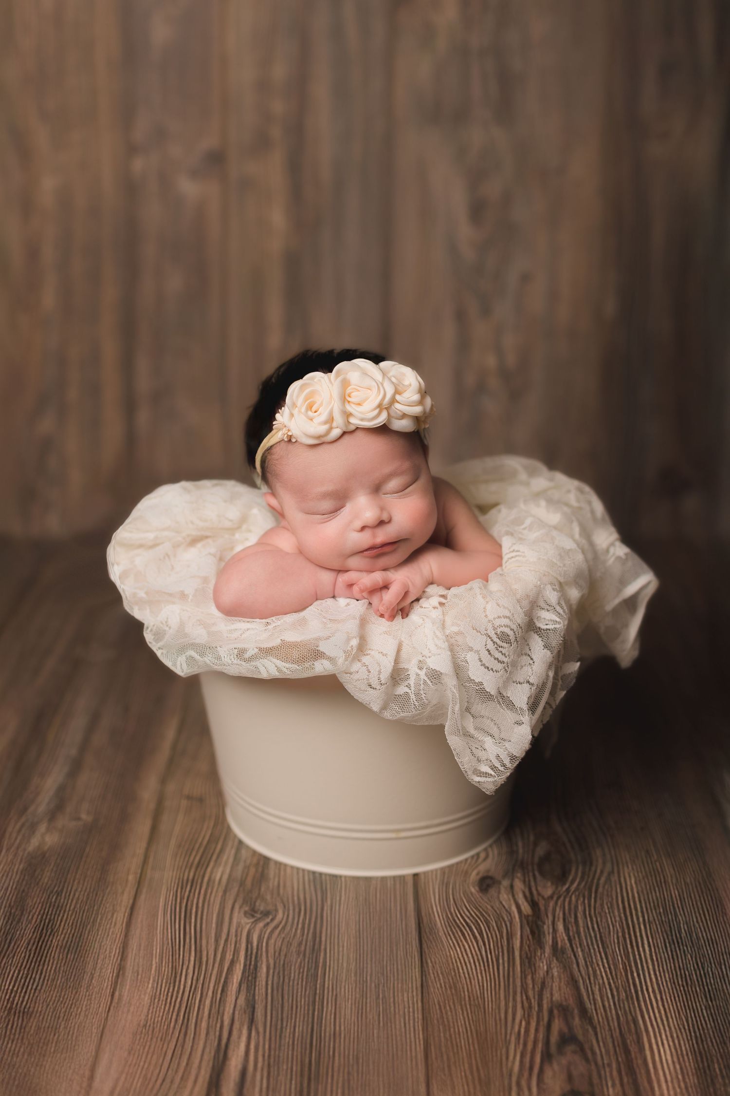 Sleeping infant in white lace wrap posed in cream-colored bowl on wooden floor.