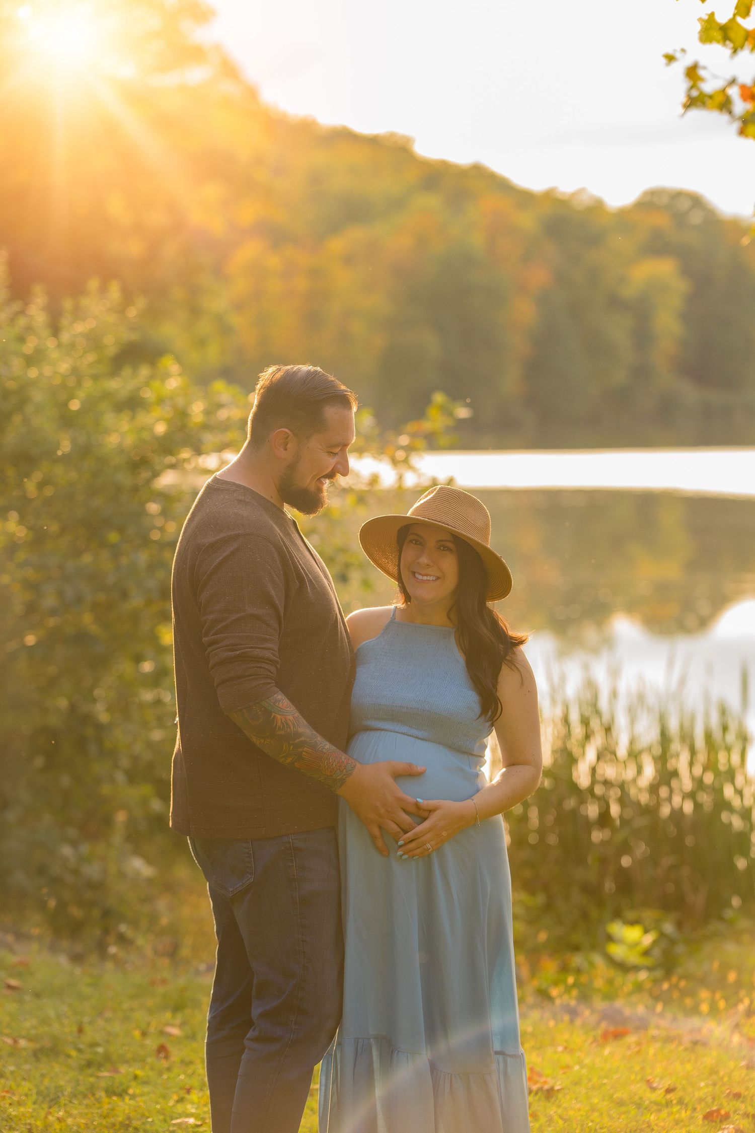 Romantic maternity photoshoot at sunset by a serene lake with couple in blue dress and dark attire.