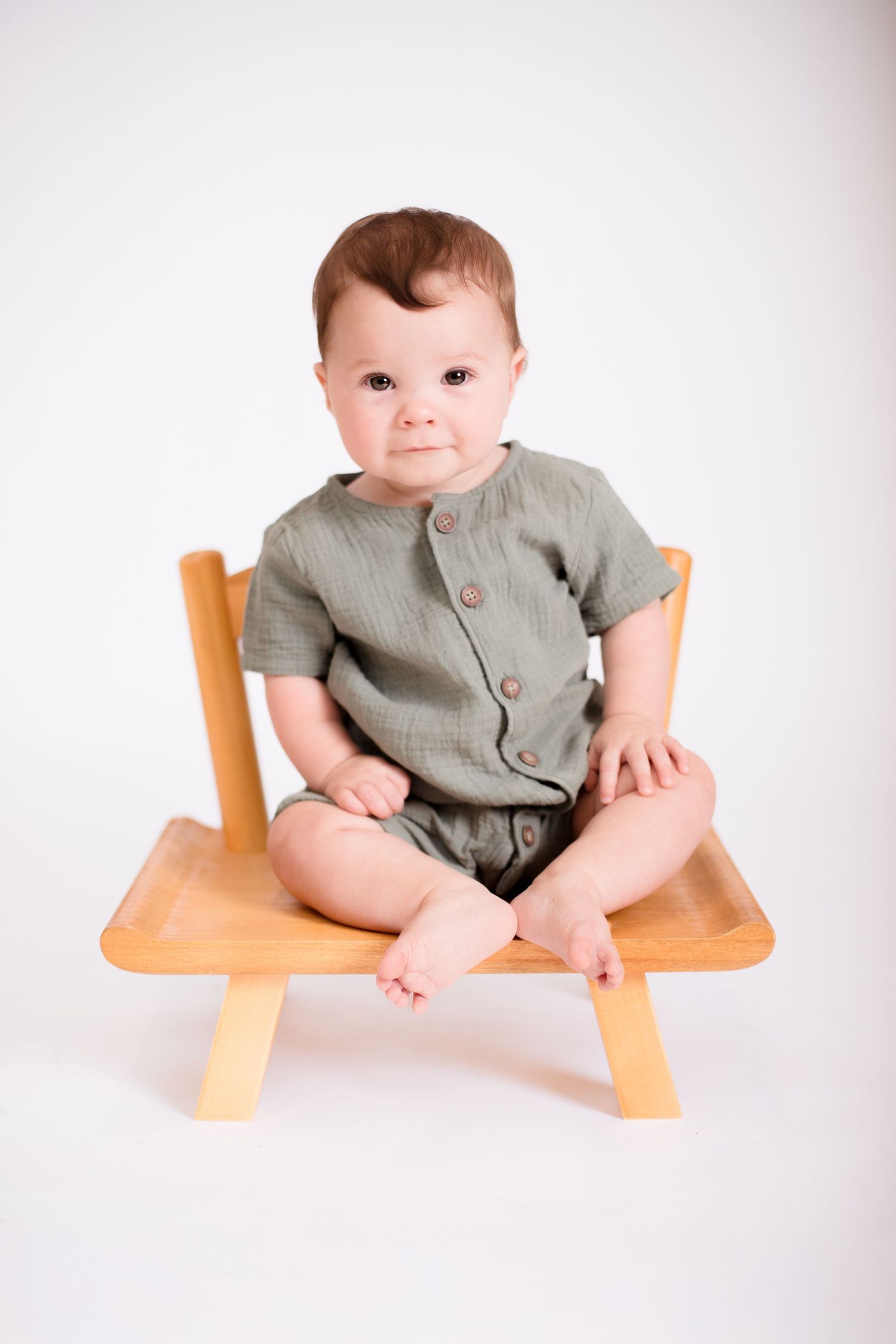 Baby in olive green outfit sits on wooden bench against white backdrop.