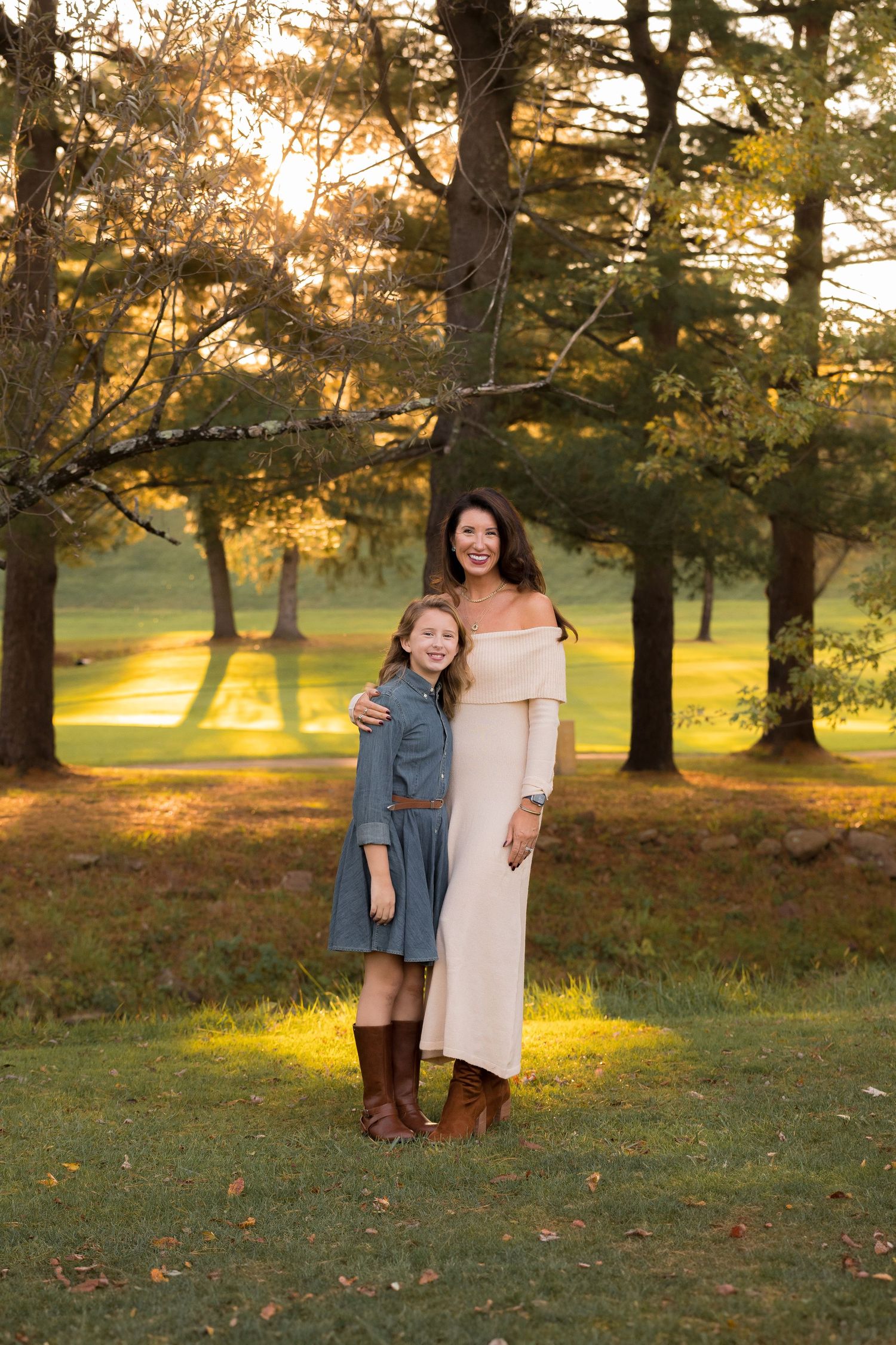 Family members stand together on grass with sunlit trees creating a warm autumn backdrop.