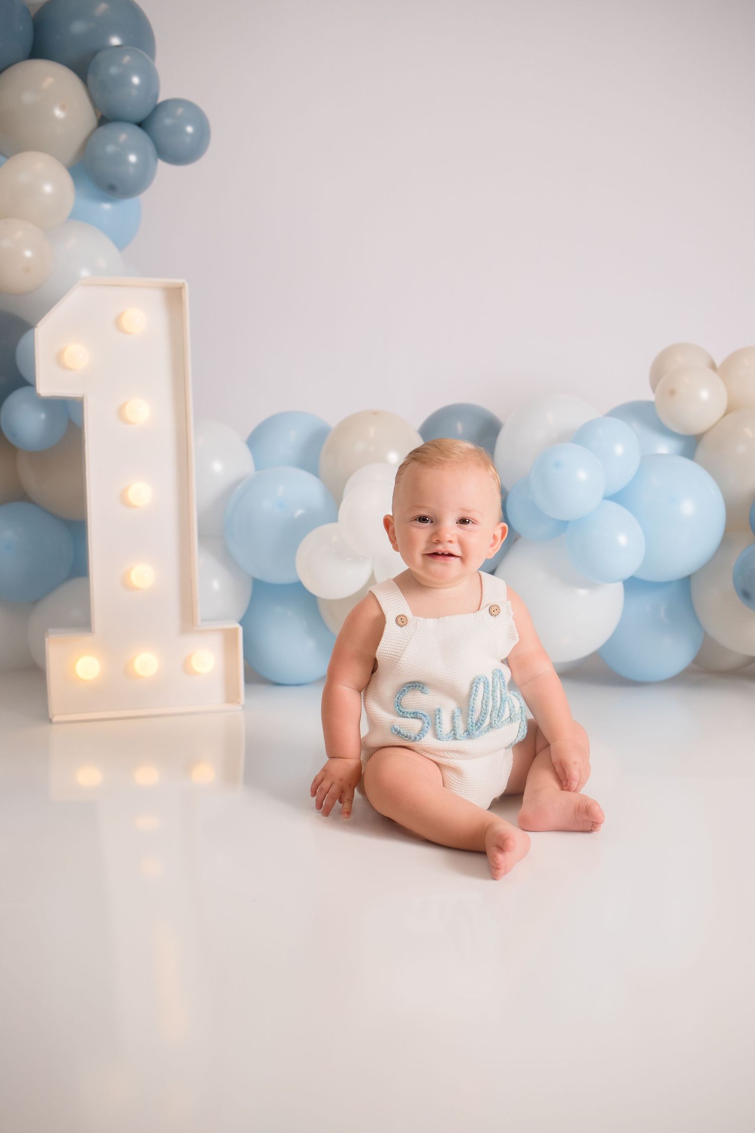 First birthday setup with illuminated number one and blue and white balloon arch.