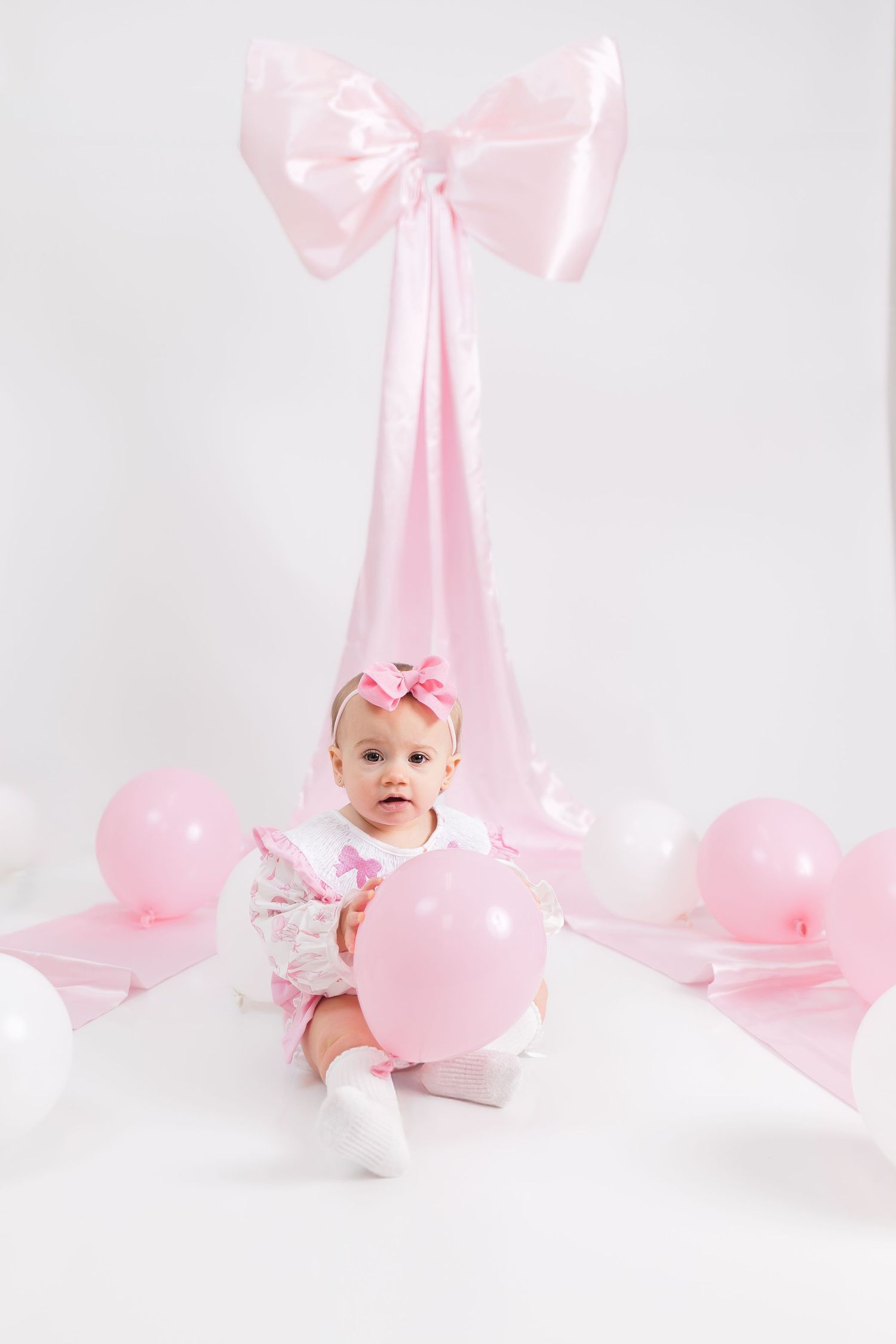 Sweet pink birthday setup with large bow backdrop, pink balloons and small baby on white floor.